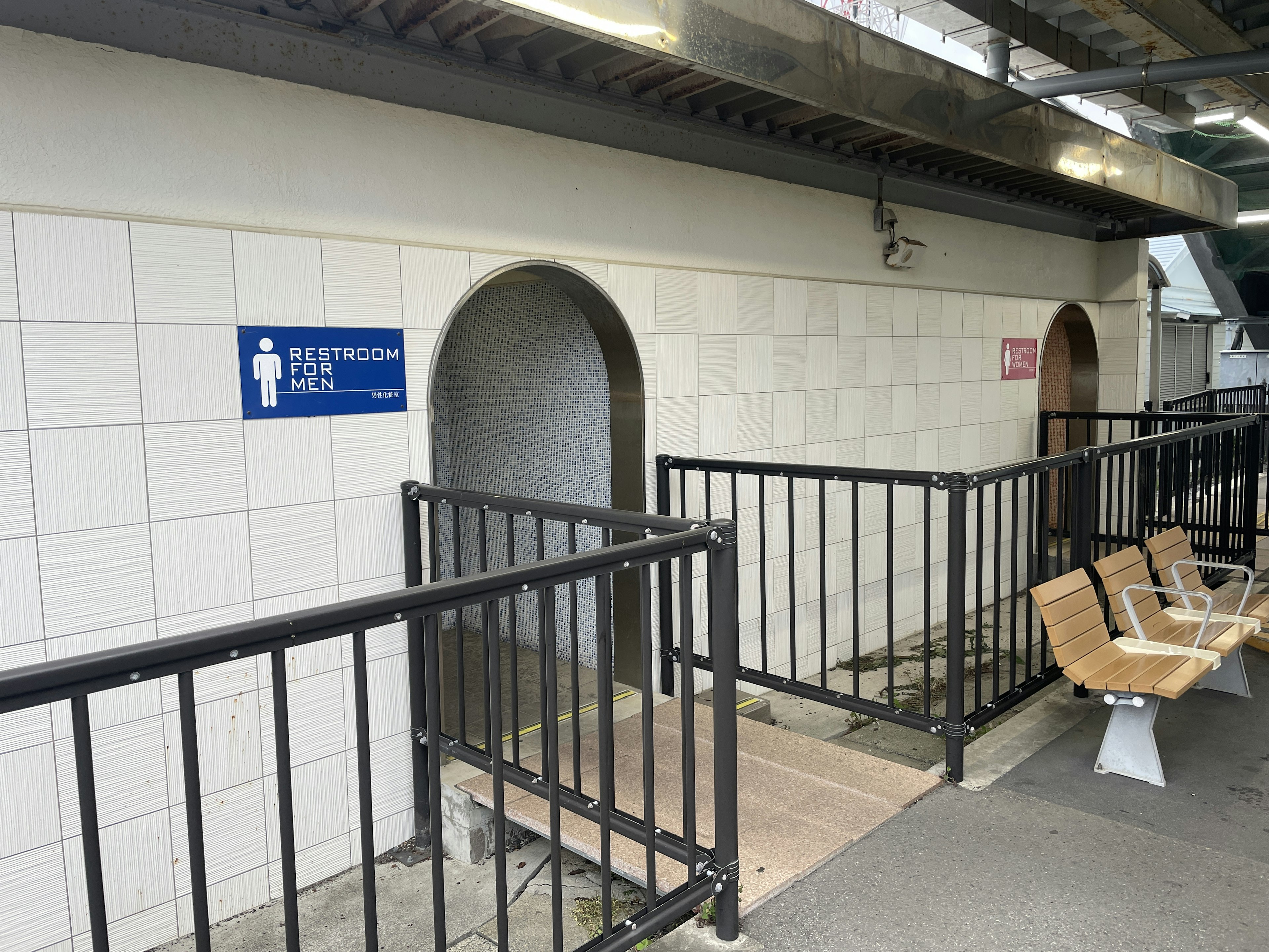 Vista exterior de un baño público con un letrero y un área cercada que incluye bancos de madera