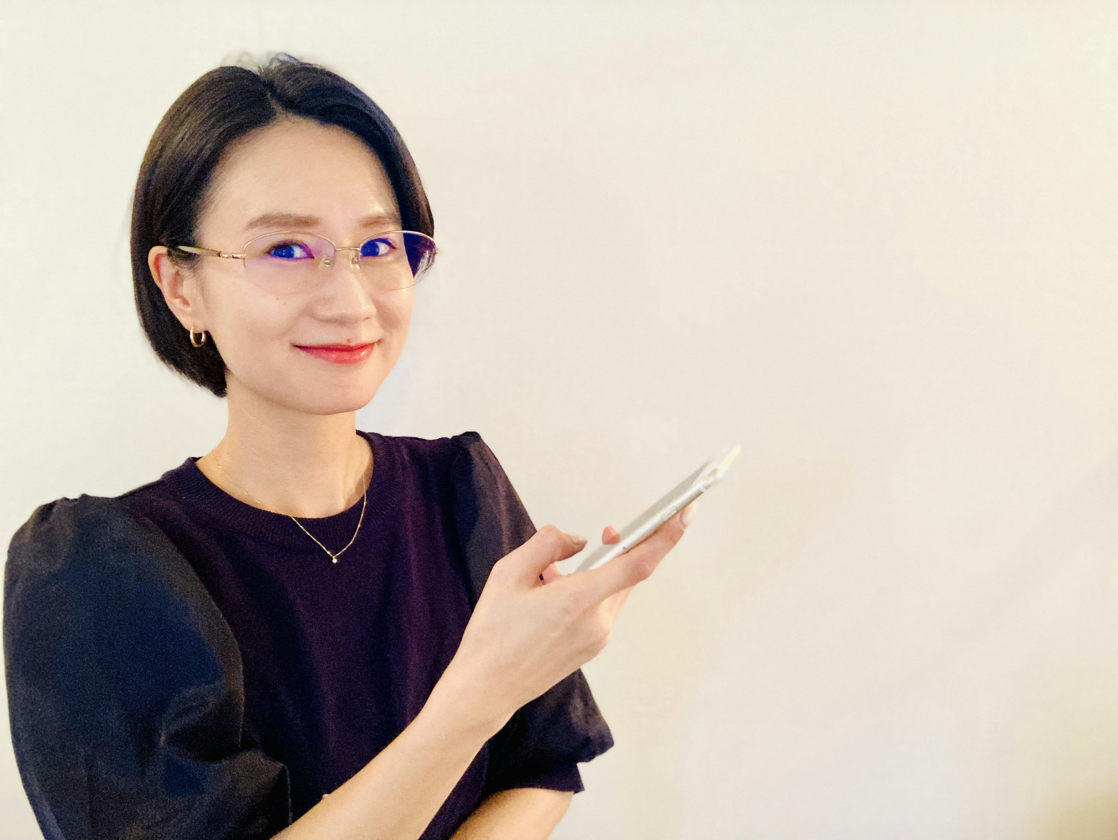 女性がスマートフォンを持ち微笑んでいる写真 短い髪型と紫の眼鏡をかけた黒いトップスを着用