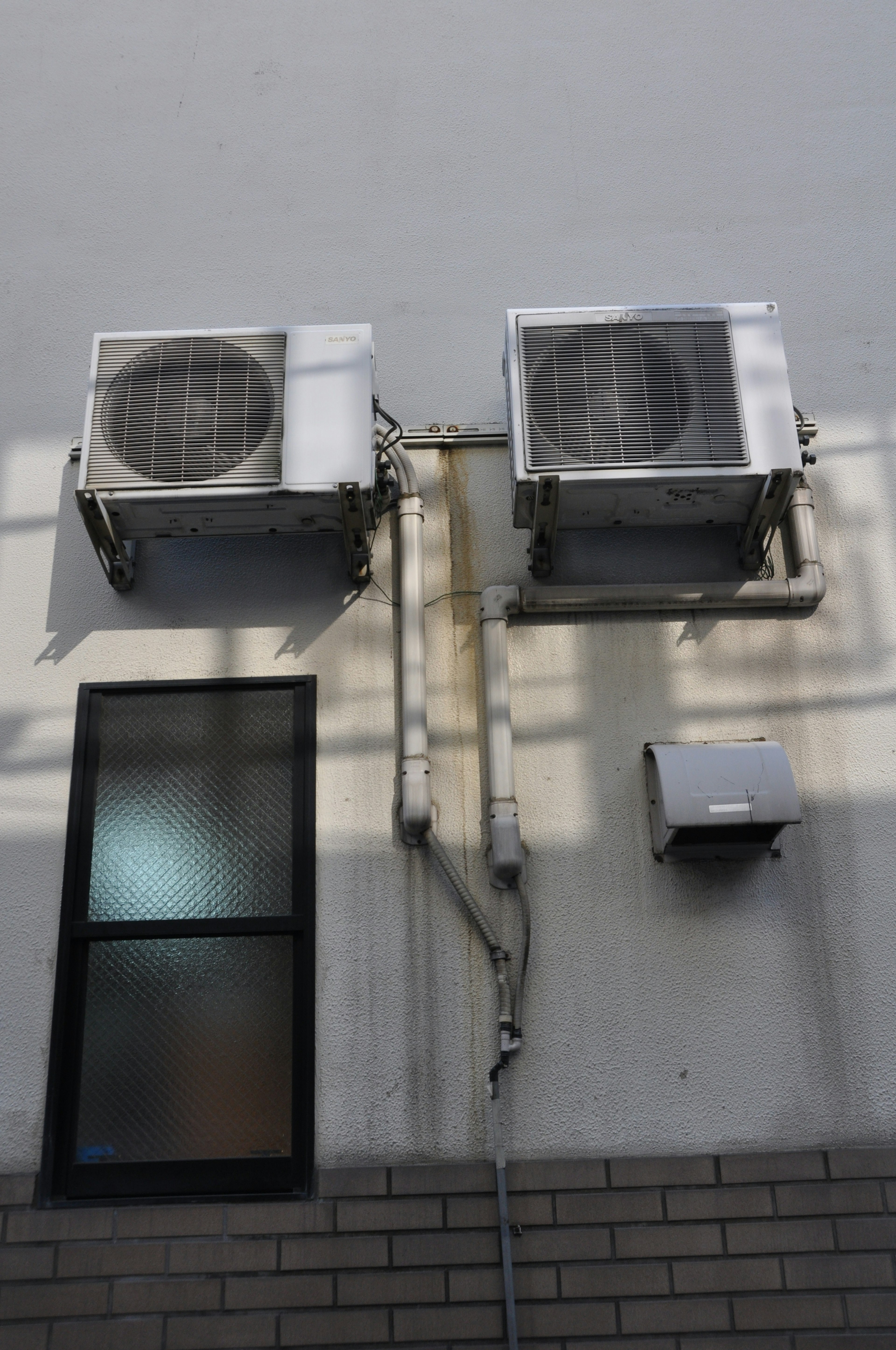 Photo of a wall with two air conditioning units installed a window and visible piping