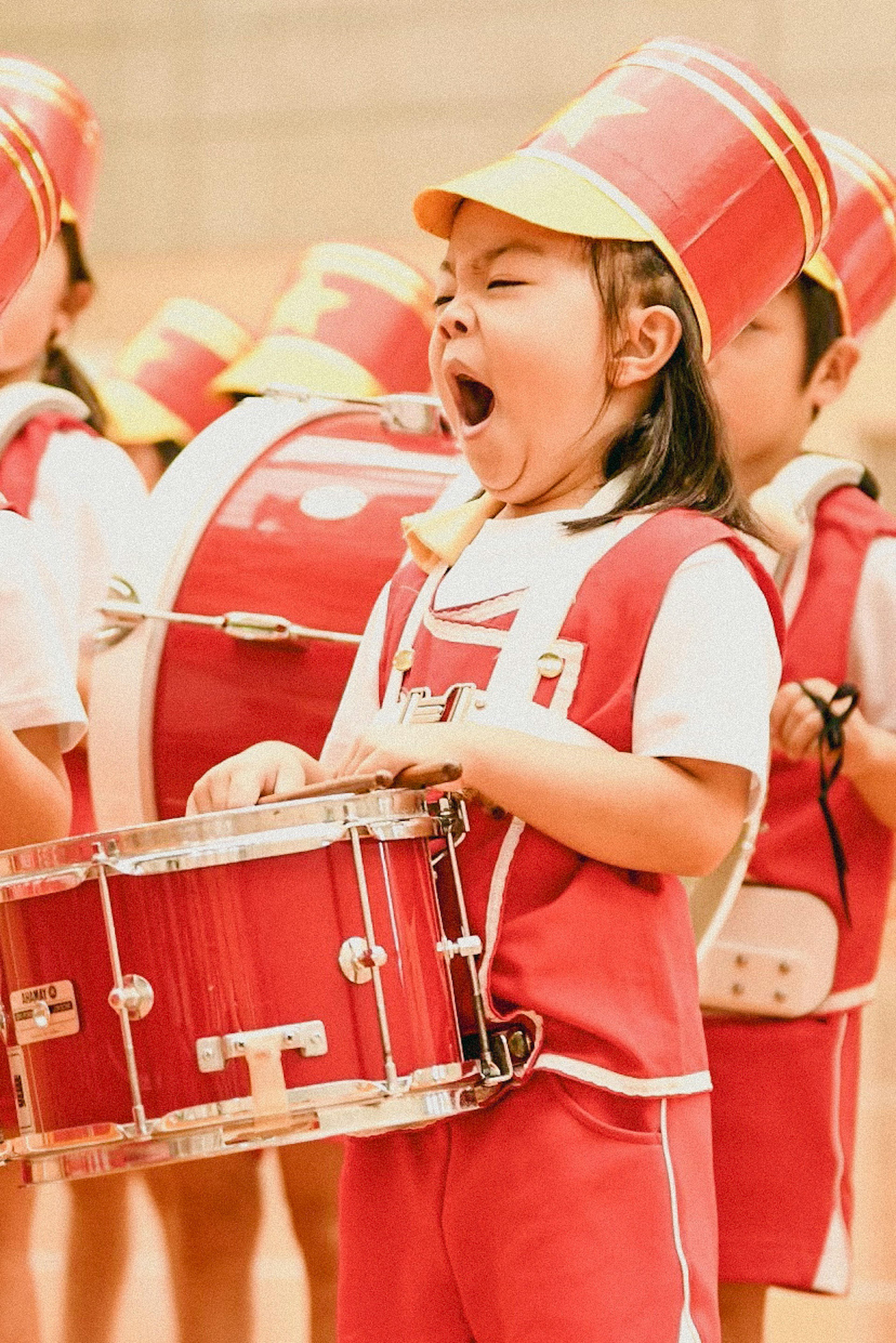 女孩在红色服装中打鼓时打哈欠，周围有其他孩子在演奏乐器