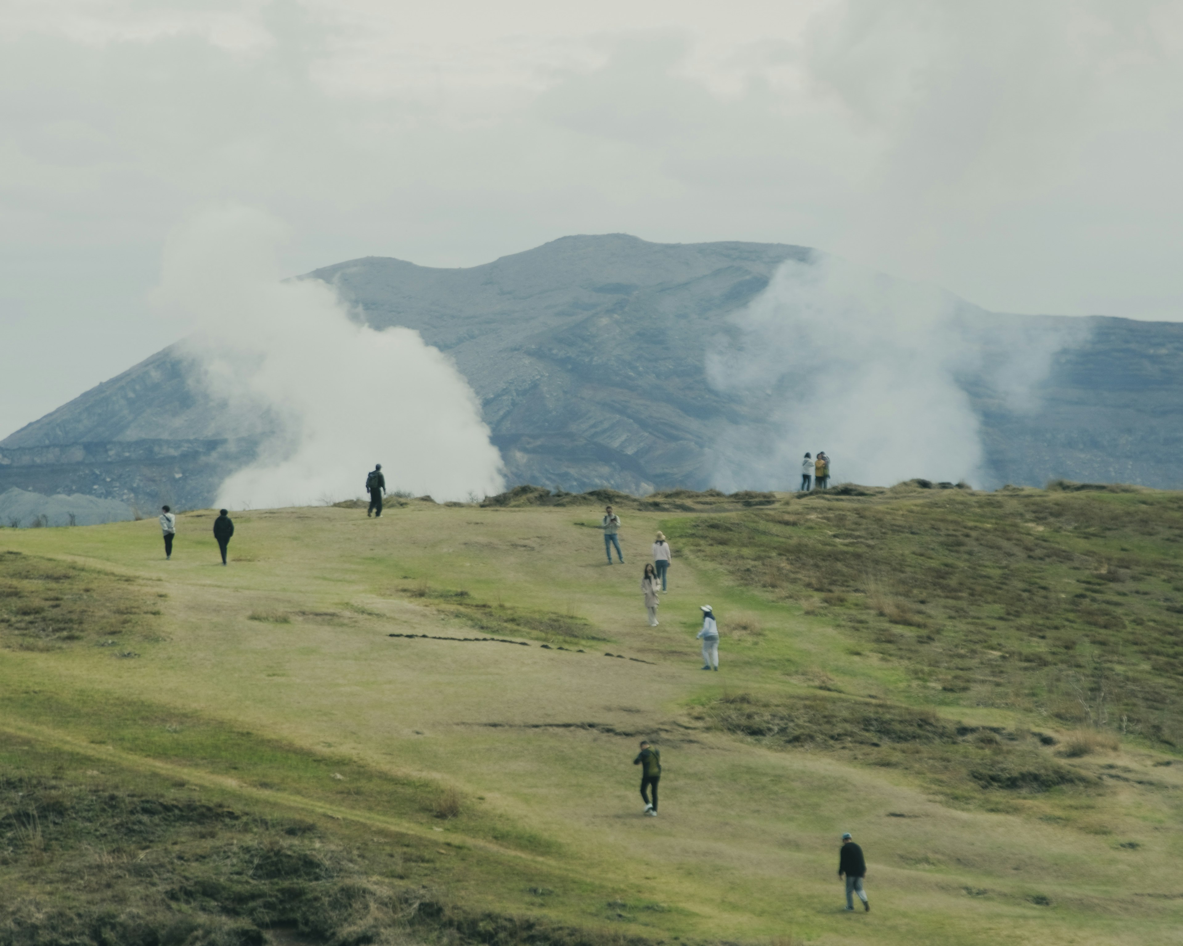 Personas caminando por una colina cubierta de hierba con humo que se eleva de las montañas al fondo