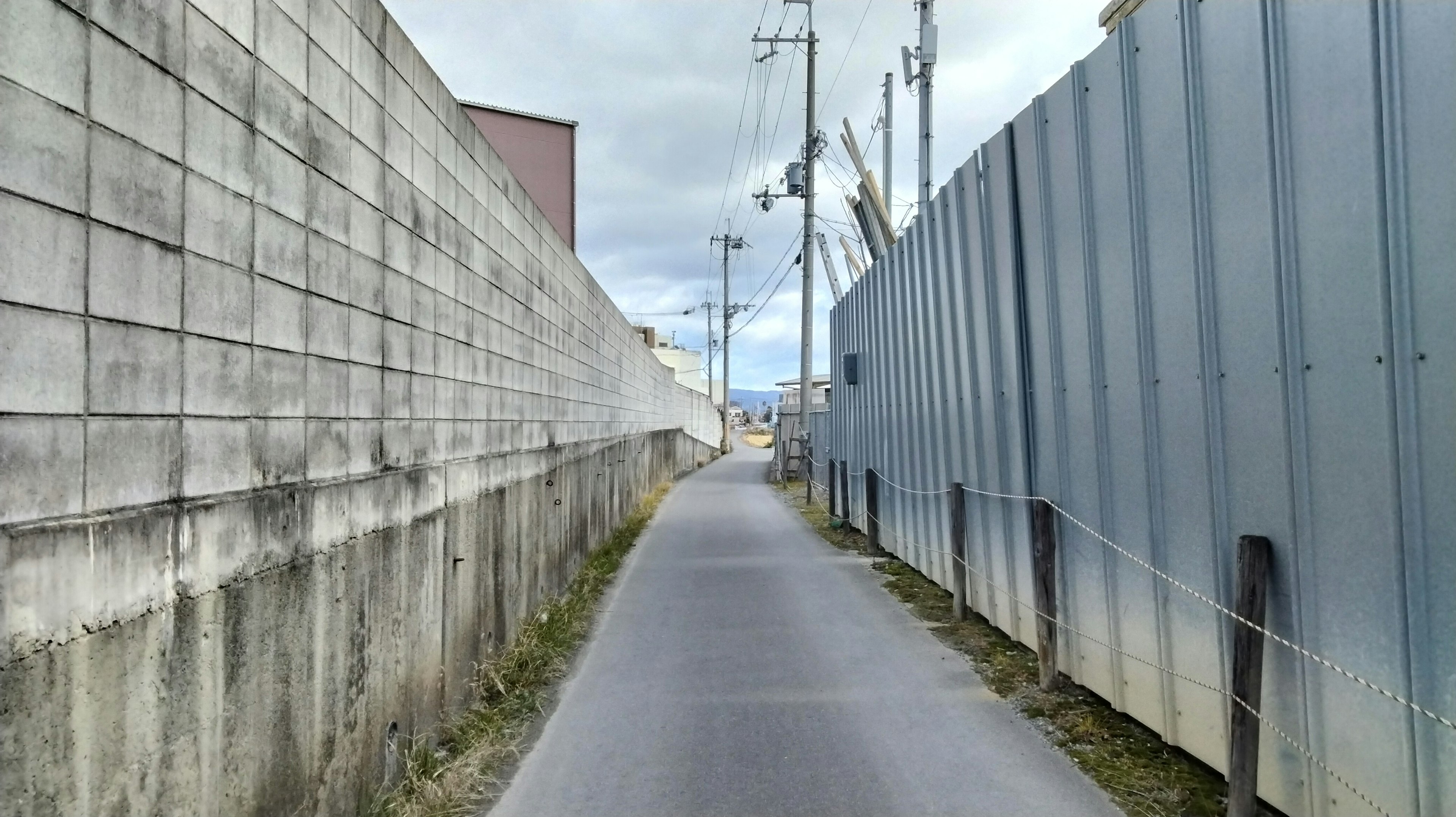 Jalan sempit yang dikelilingi oleh dinding beton dan pagar logam di bawah langit mendung