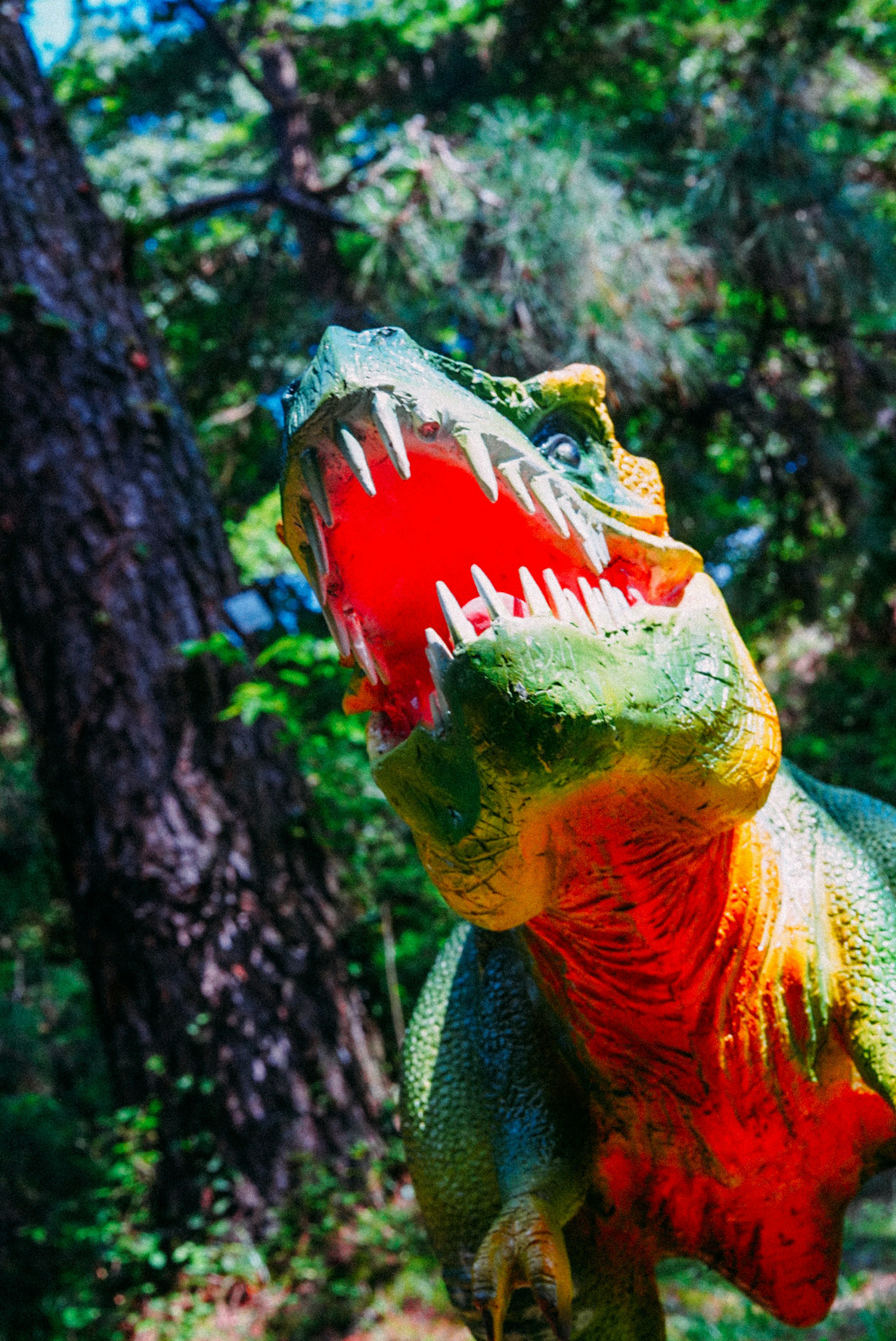 A green dinosaur model roaring with its mouth open surrounded by trees