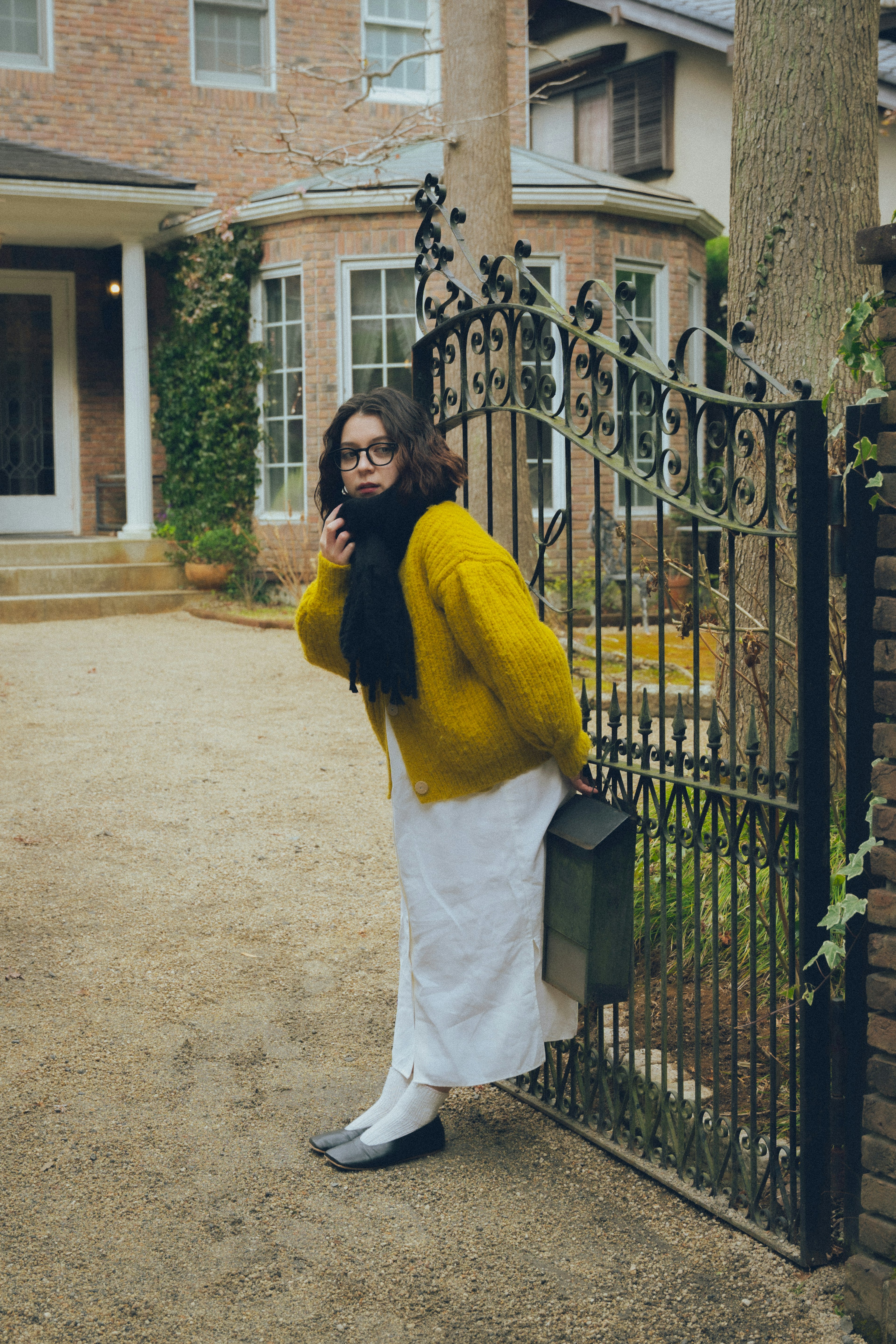 A woman in a yellow sweater and white skirt with a black scarf standing by a gate