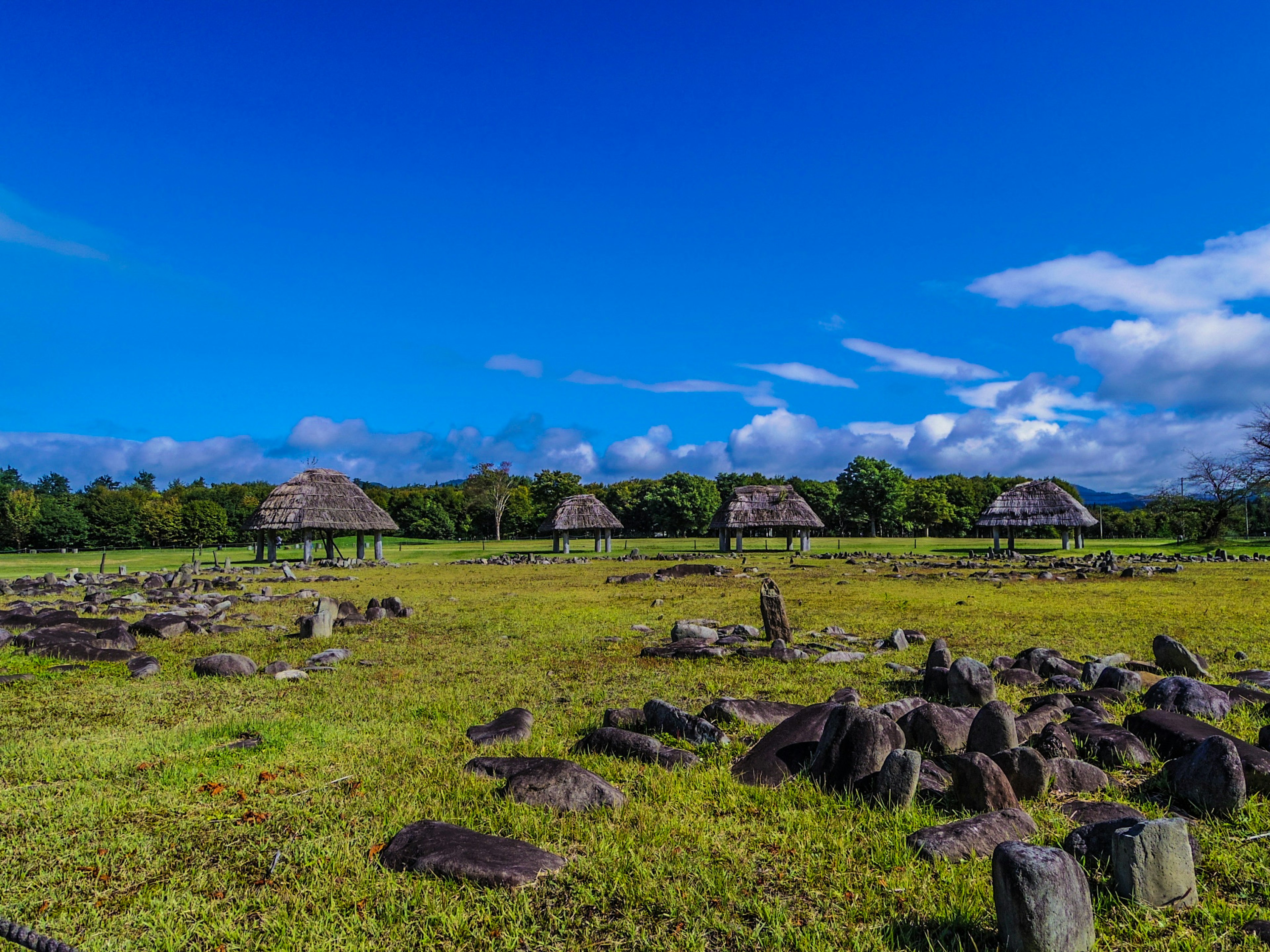 A landscape with huts on a grassy field under a blue sky ancient stone ruins scattered throughout