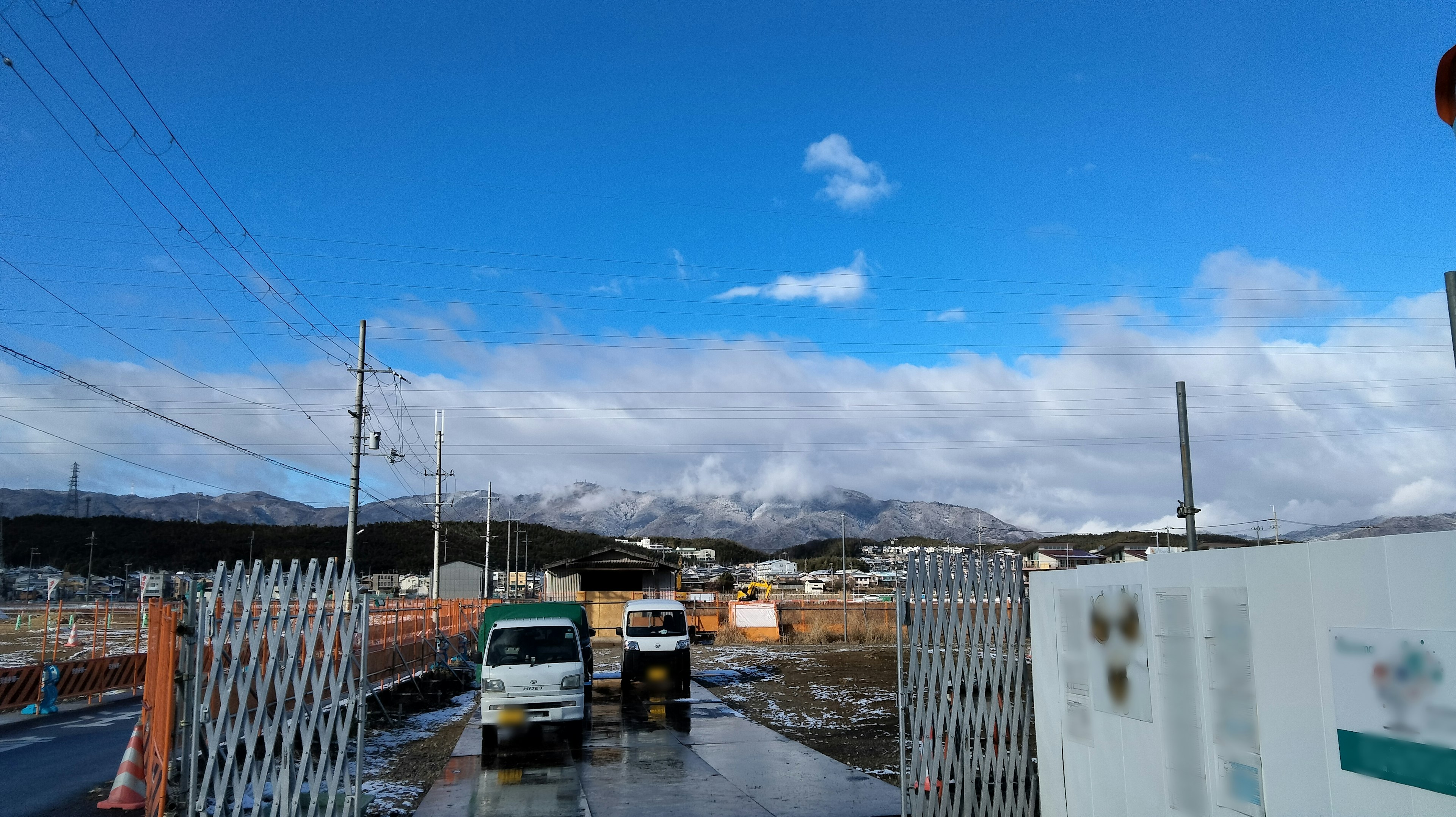 晴れた空と雲が広がる風景に、山々が背景に見える建設現場の入り口