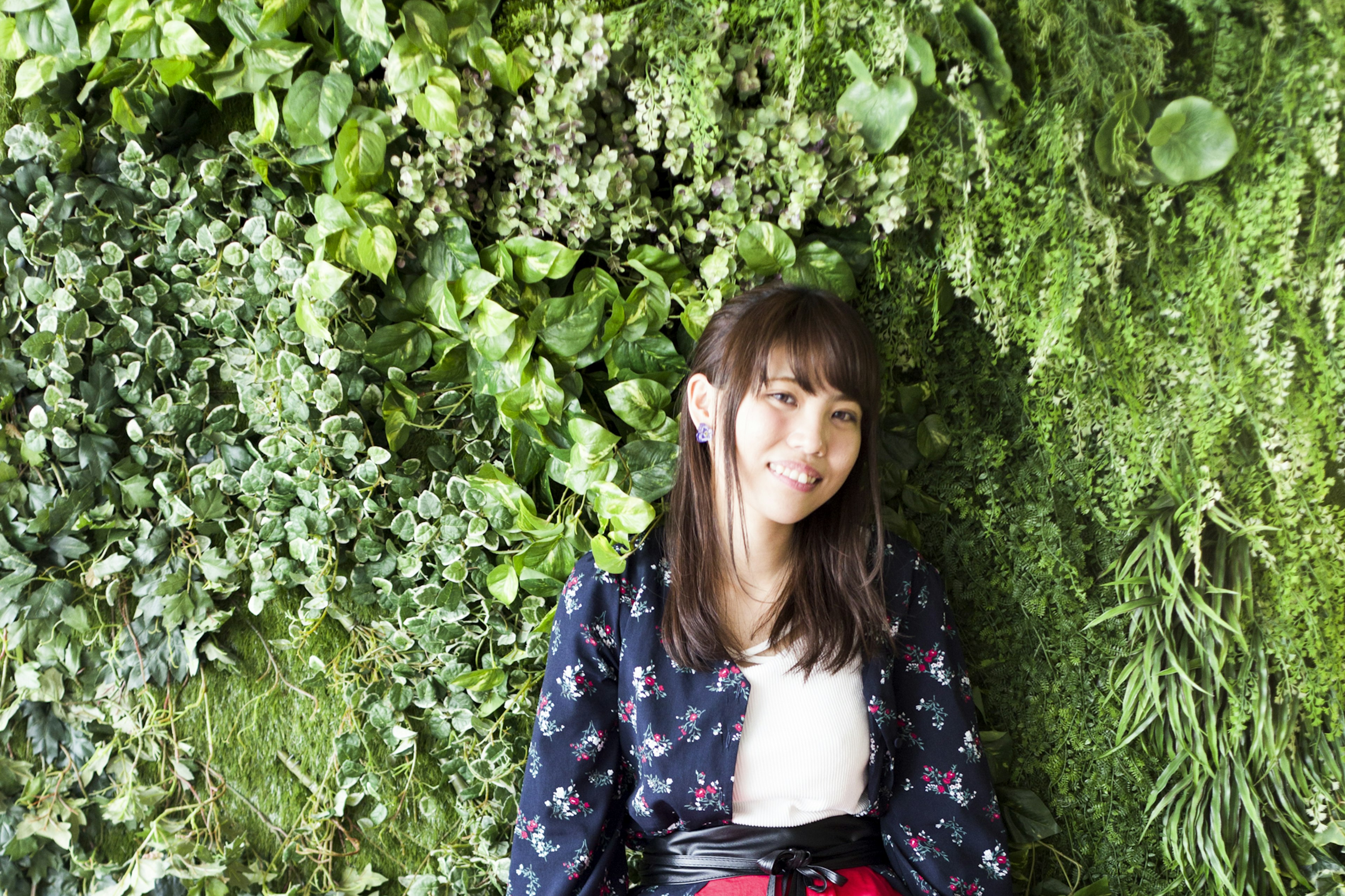 Donna seduta di fronte a un muro verde lussureggiante Sorridente e che guarda la fotocamera Indossa una giacca floreale