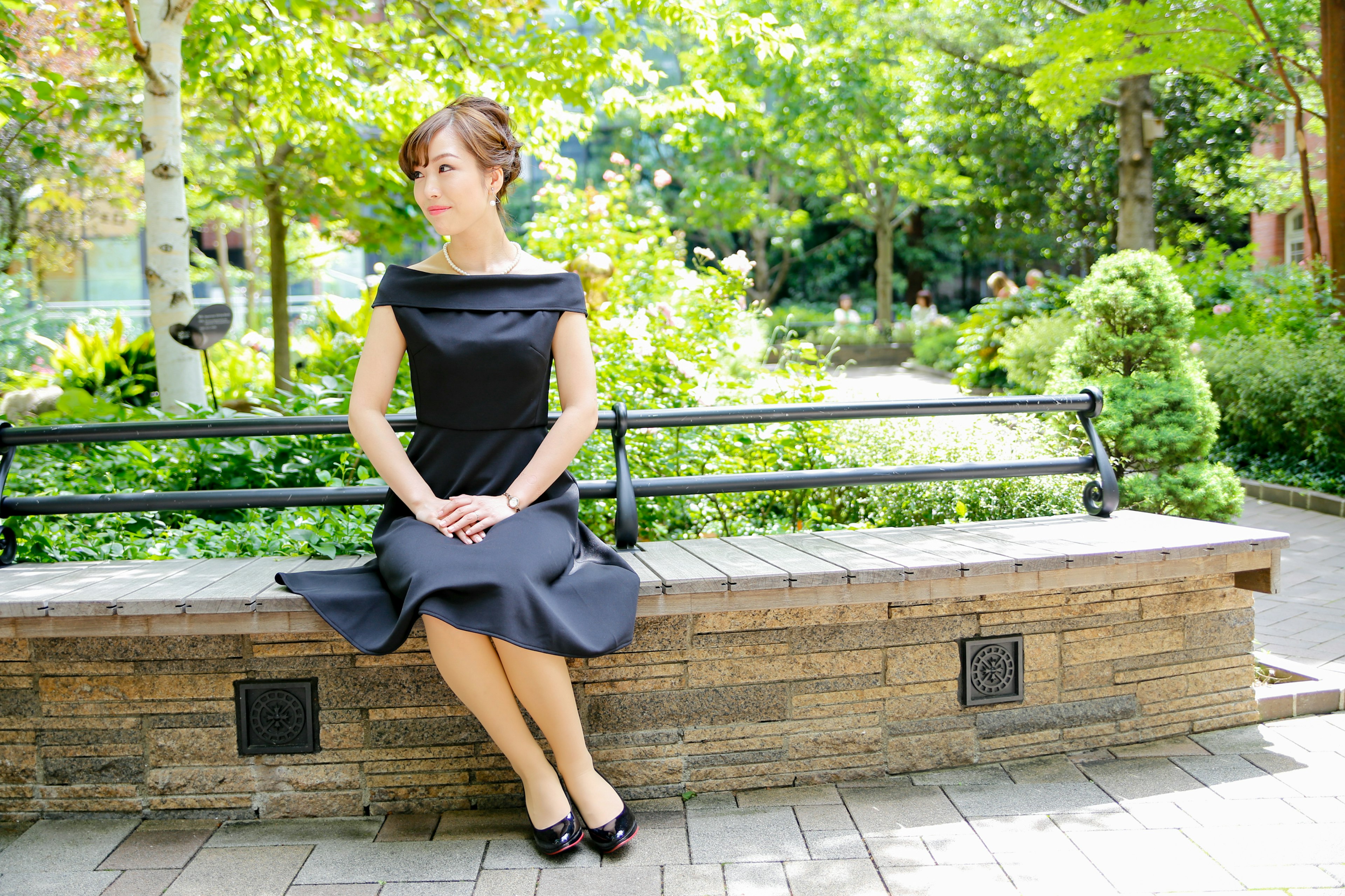 A woman sitting on a park bench wearing a black dress surrounded by green trees