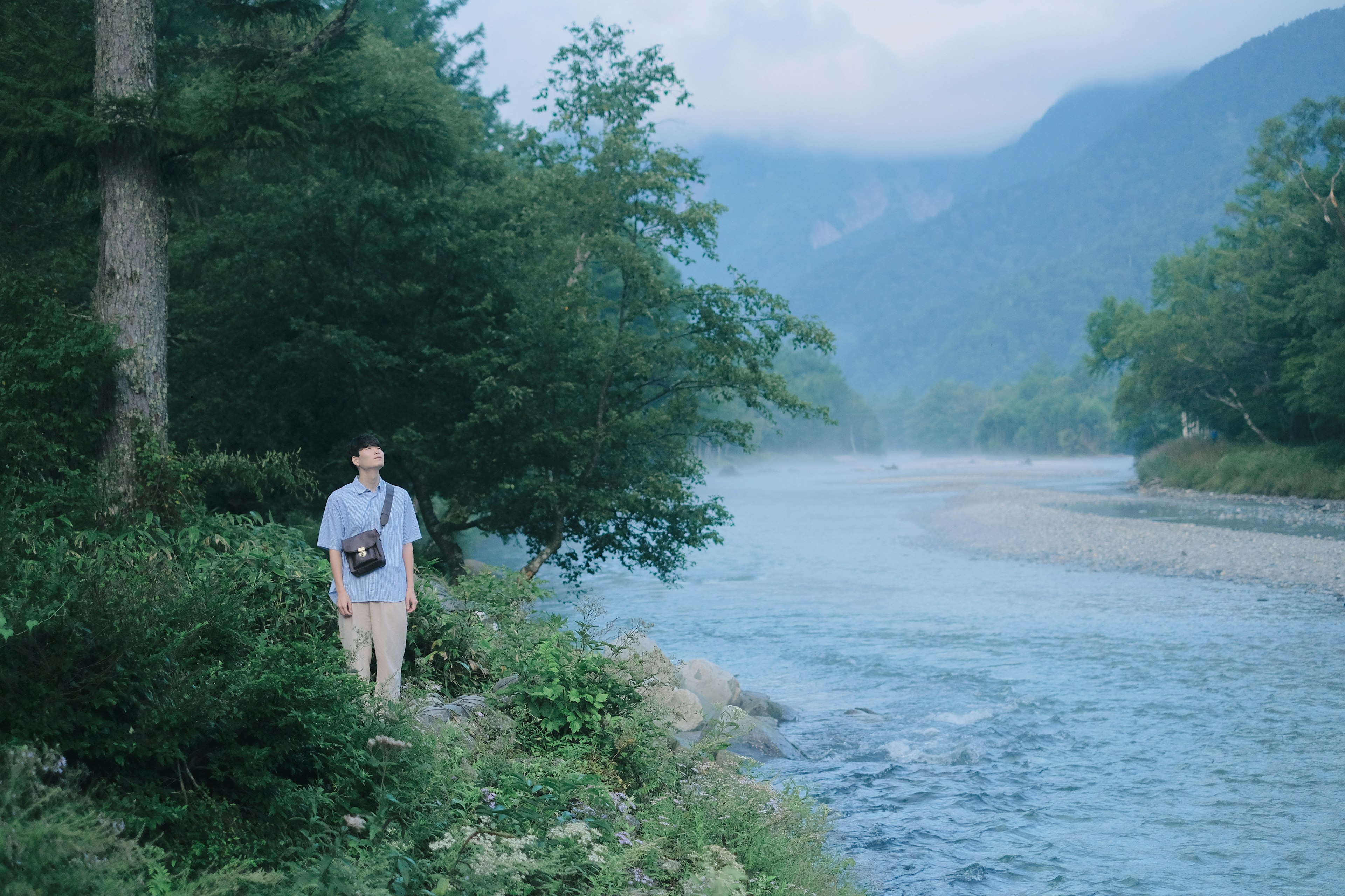 Persona in piedi vicino a un fiume blu circondata da alberi verdi e montagne