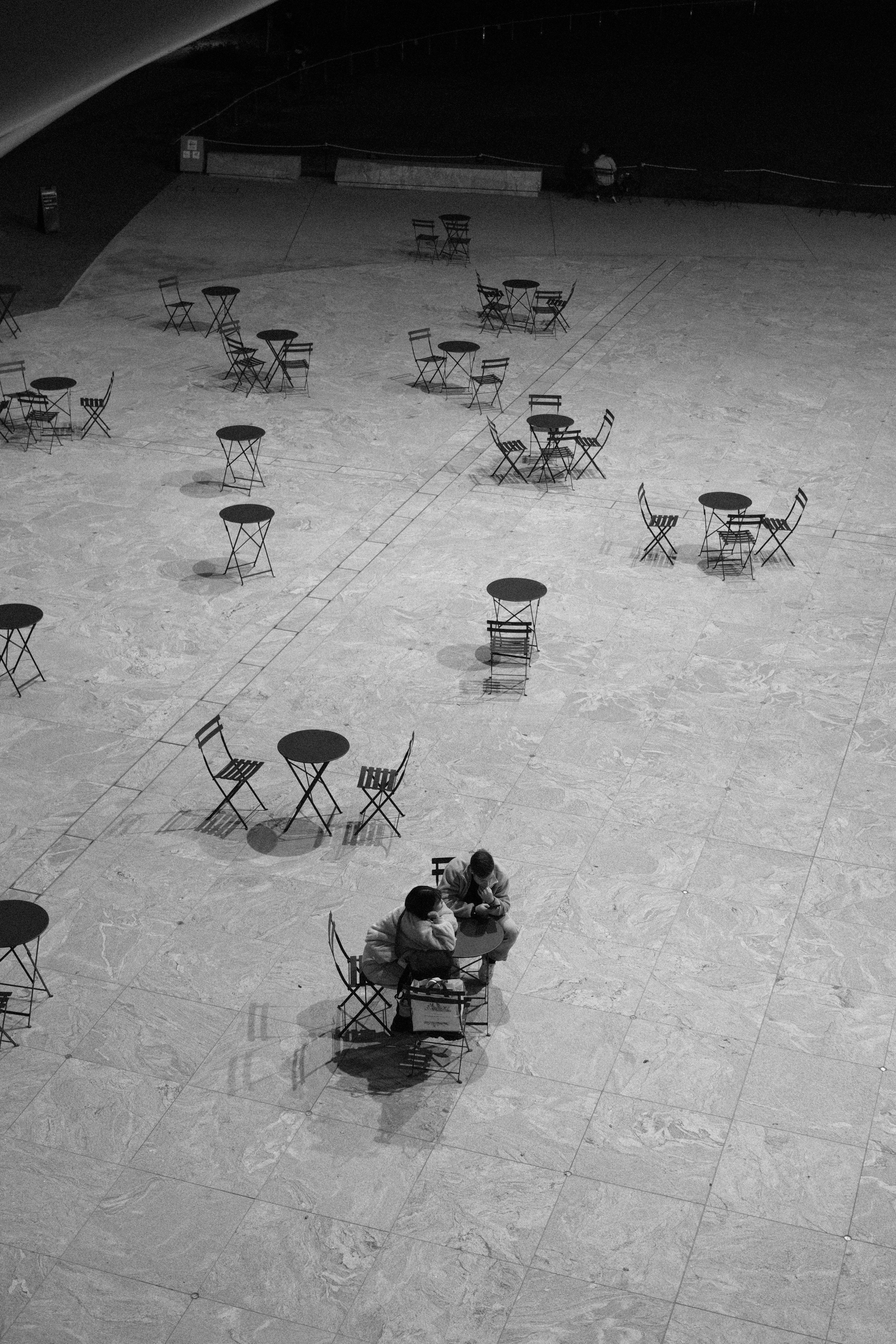 A monochrome image of two people sitting at a table in a spacious area filled with scattered tables and chairs