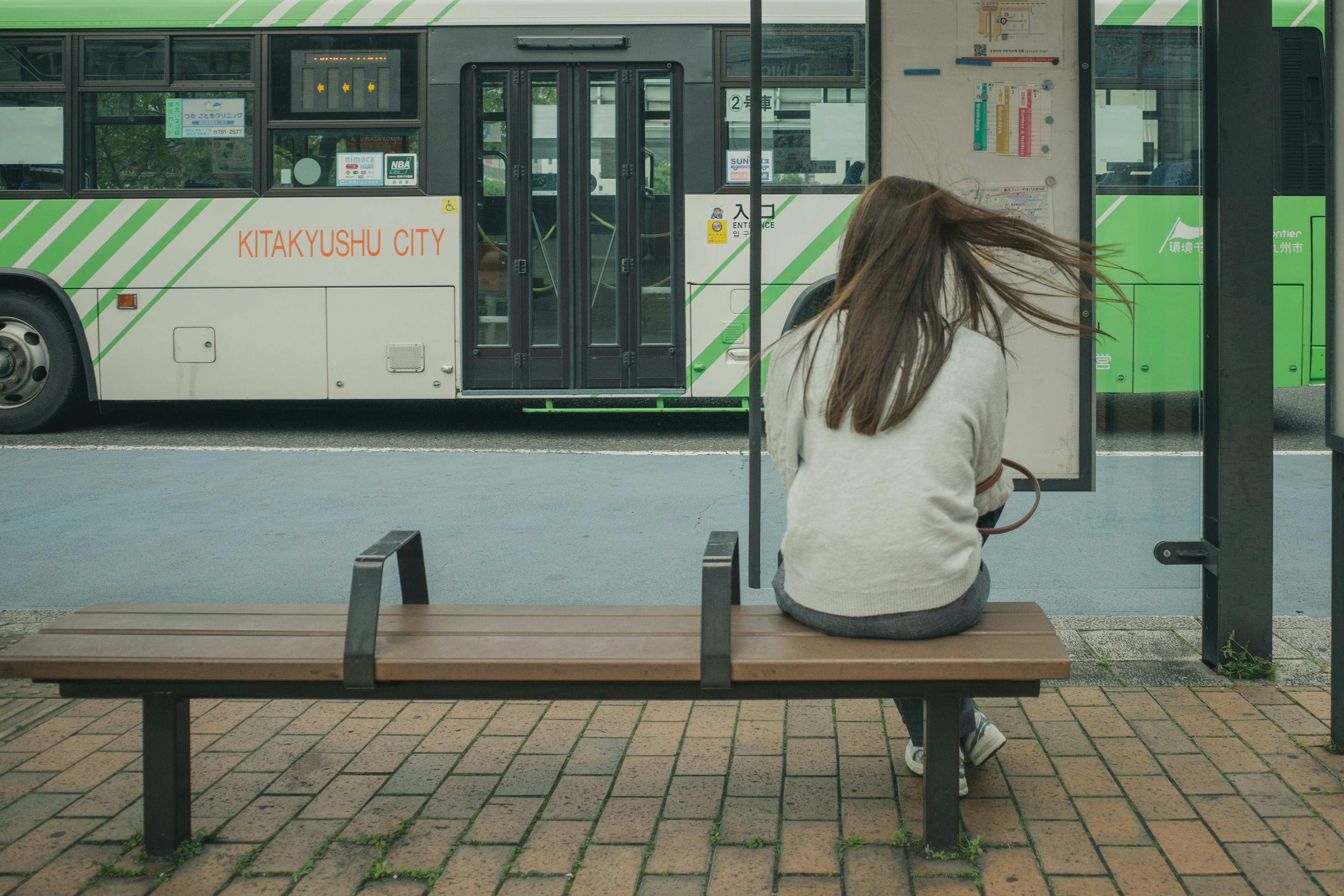 A woman sitting at a bus stop with flowing hair and a bus in the background