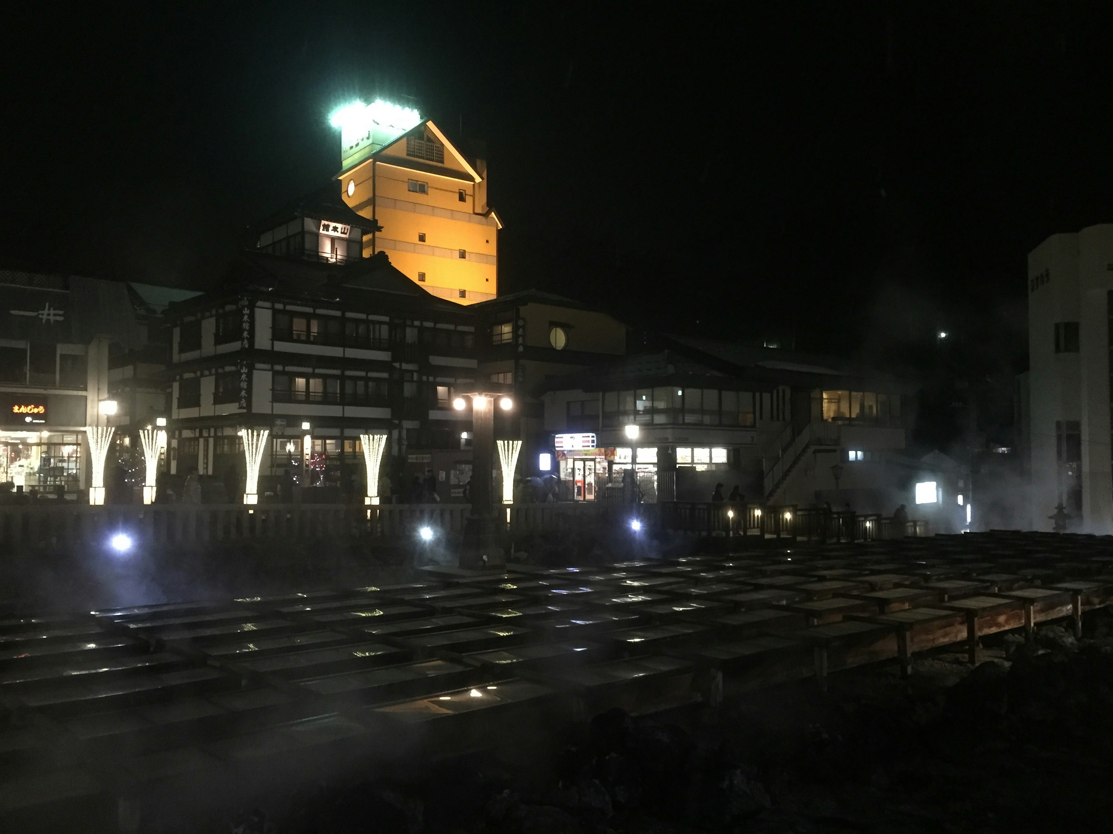 夜の温泉街の風景で、照明が美しい建物と蒸気が立ち上る温泉が見える