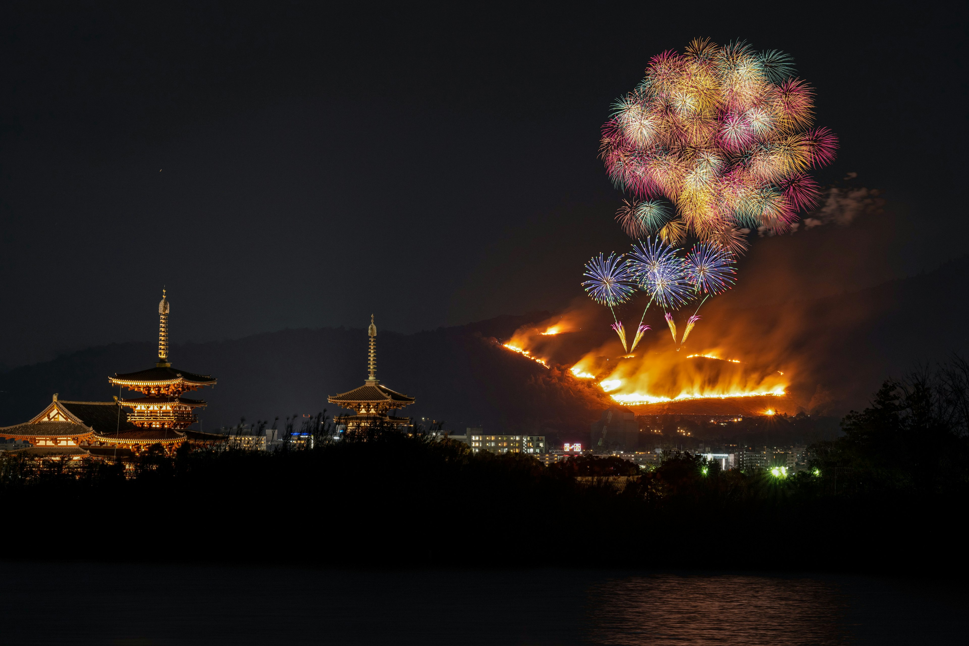 Fireworks illuminate the night sky above a mountain with flames