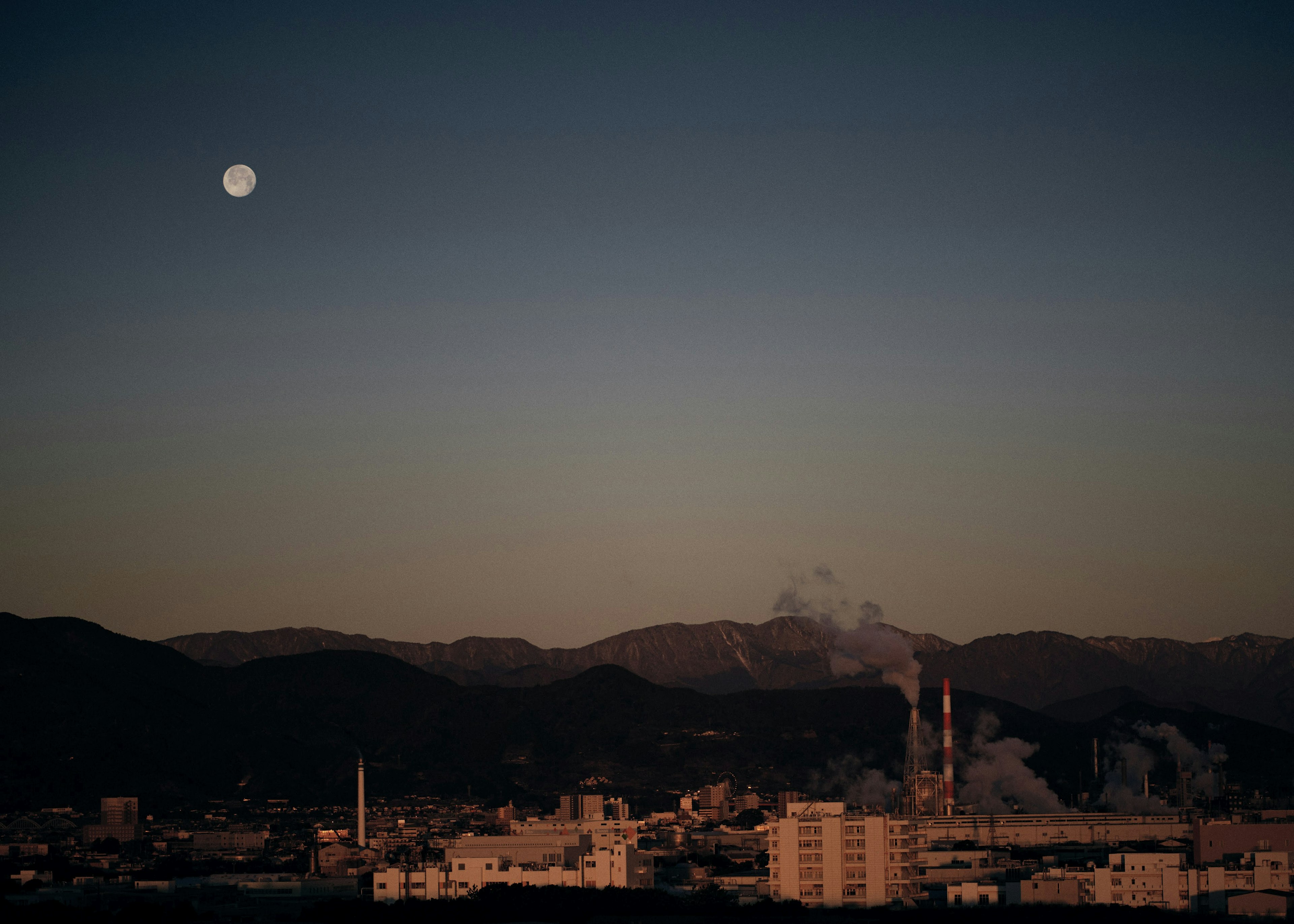 夜空に浮かぶ満月と工場の煙が見える山の風景