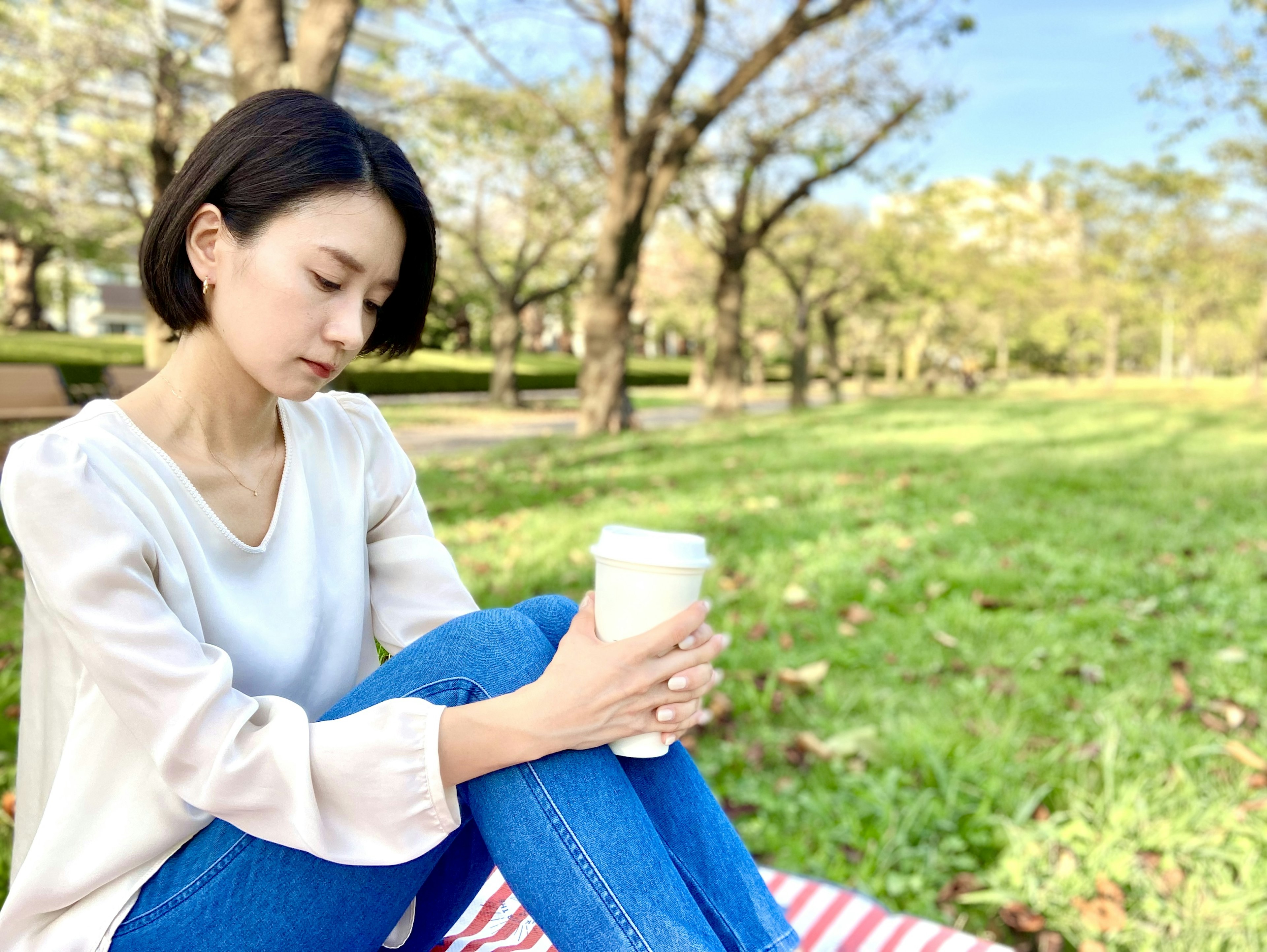 公園でコーヒーを持って座っている女性　緑の芝生と木々が背景