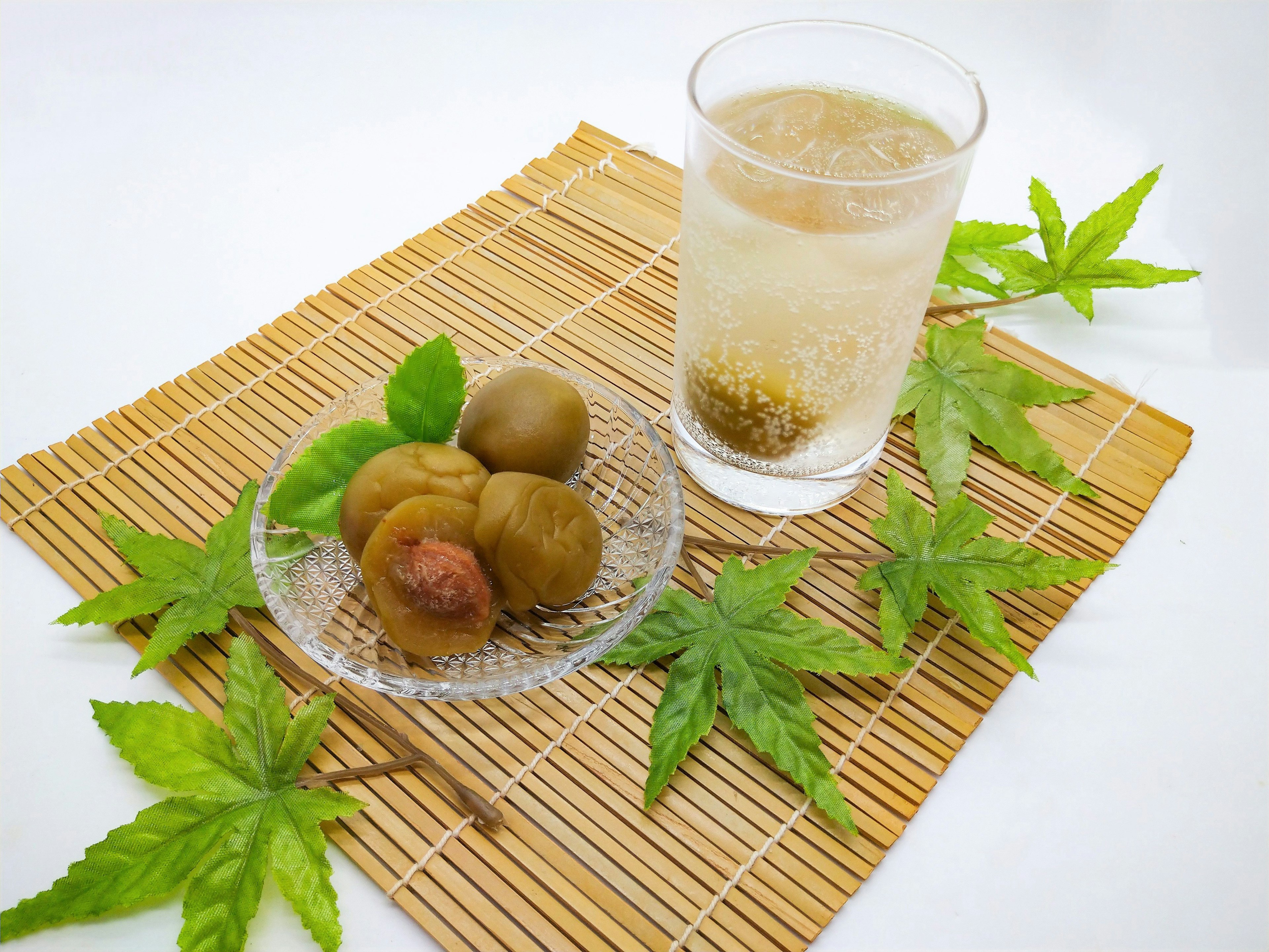 A bamboo mat with a glass of drink and a plate of pickled plums surrounded by green leaves