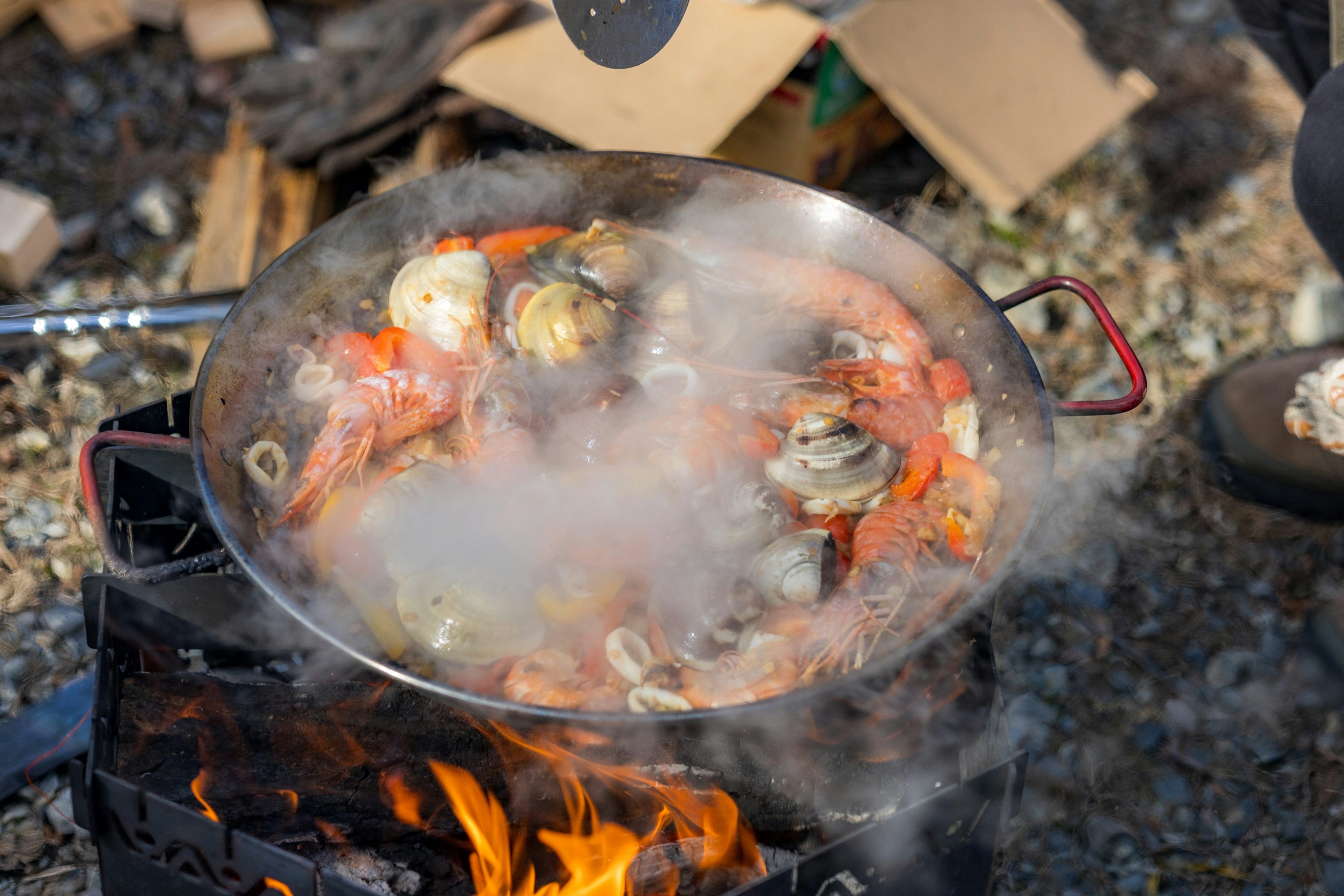 A pot of seafood paella cooking over an open flame steam rising colorful ingredients visible