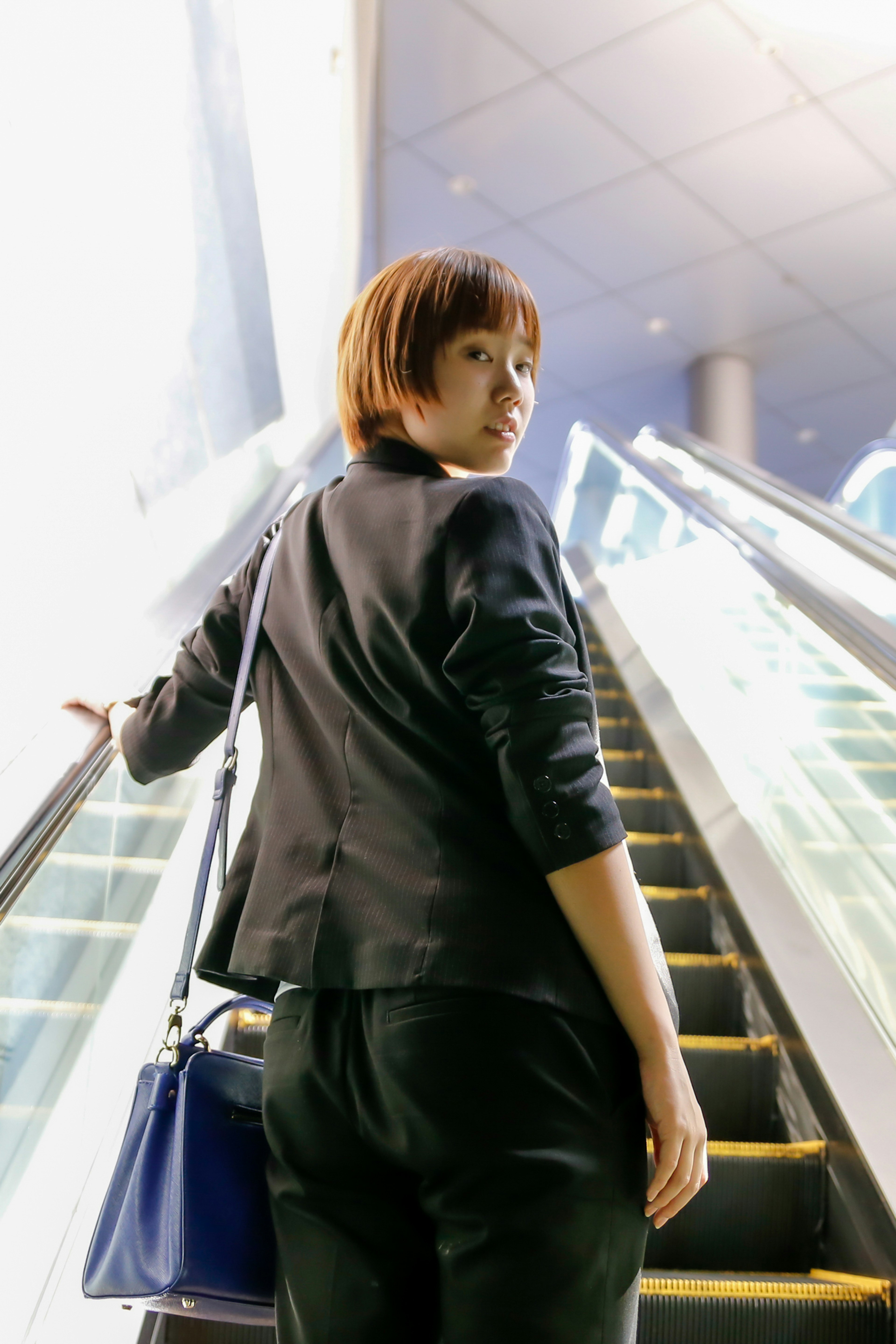 Woman ascending an escalator viewed from behind Short hair black jacket blue bag