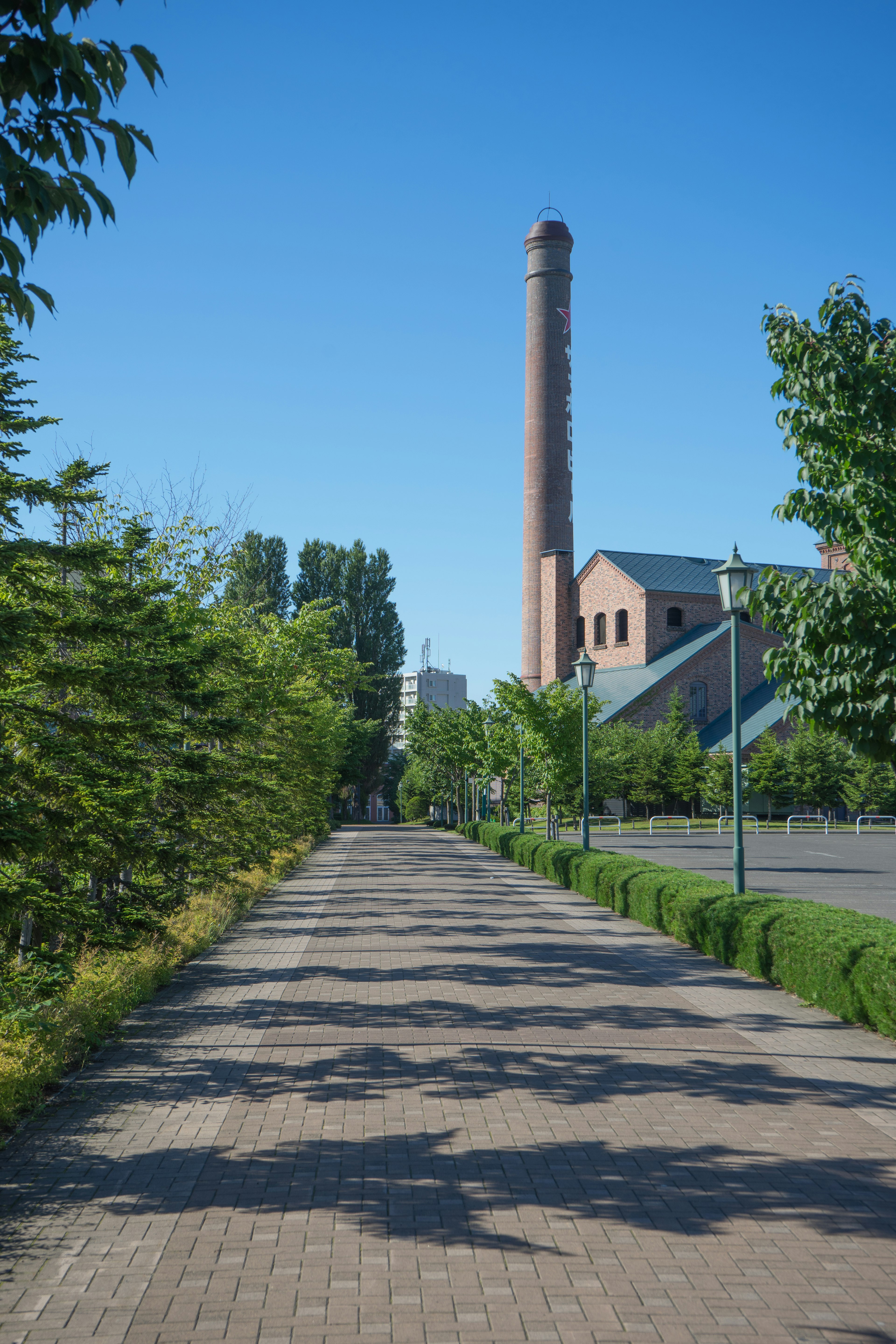 Allée pavée bordée d'arbres verts et d'une cheminée d'usine sous un ciel bleu