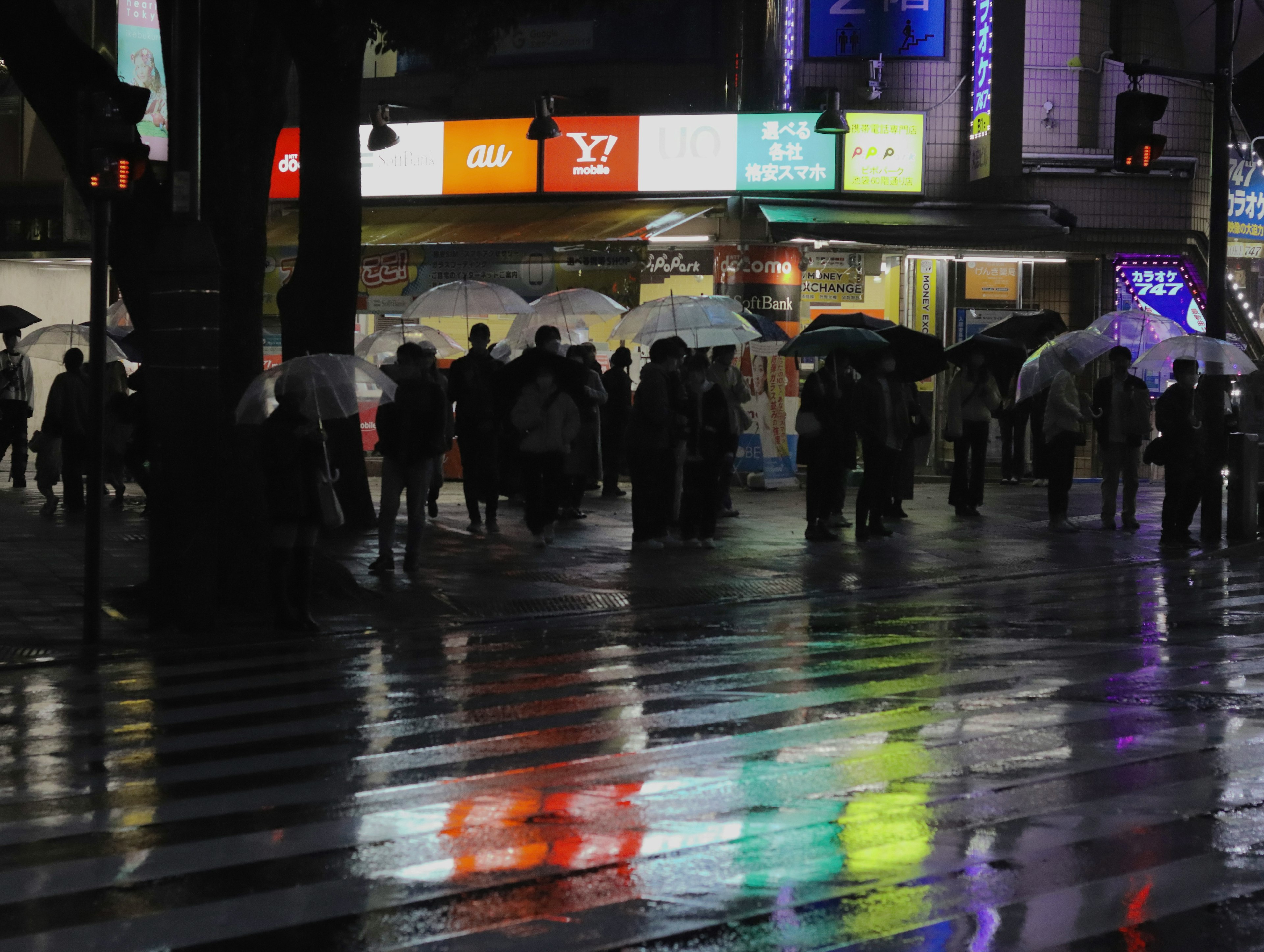 Personas con paraguas en un cruce reflejando luces coloridas por la noche