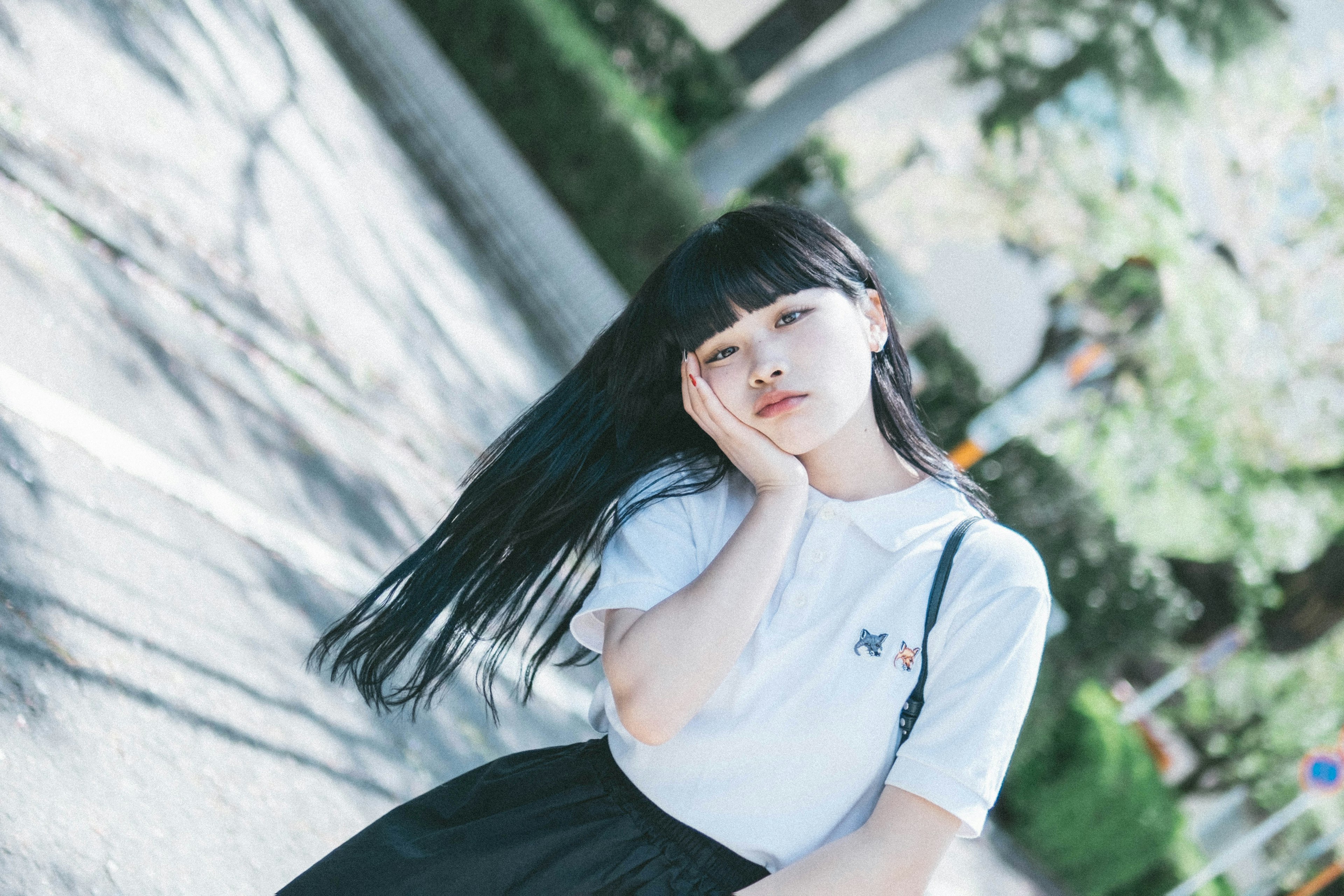 Une fille aux longs cheveux noirs posant dans la rue, portant un t-shirt blanc et une jupe noire, la main sur la joue