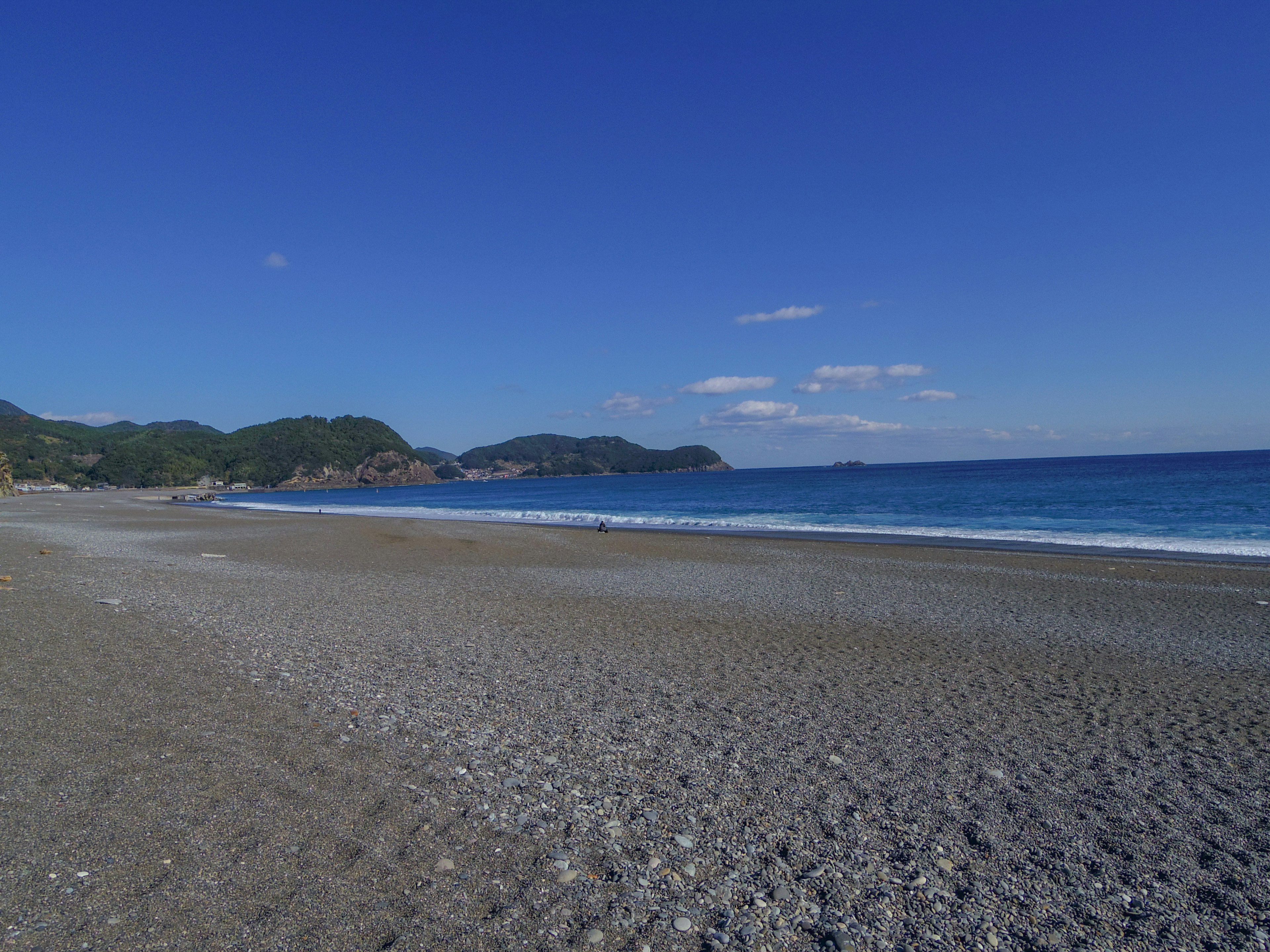 Malerscher Ausblick auf einen blauen Himmel und Meer mit einem Kiesstrand und grünen Hügeln