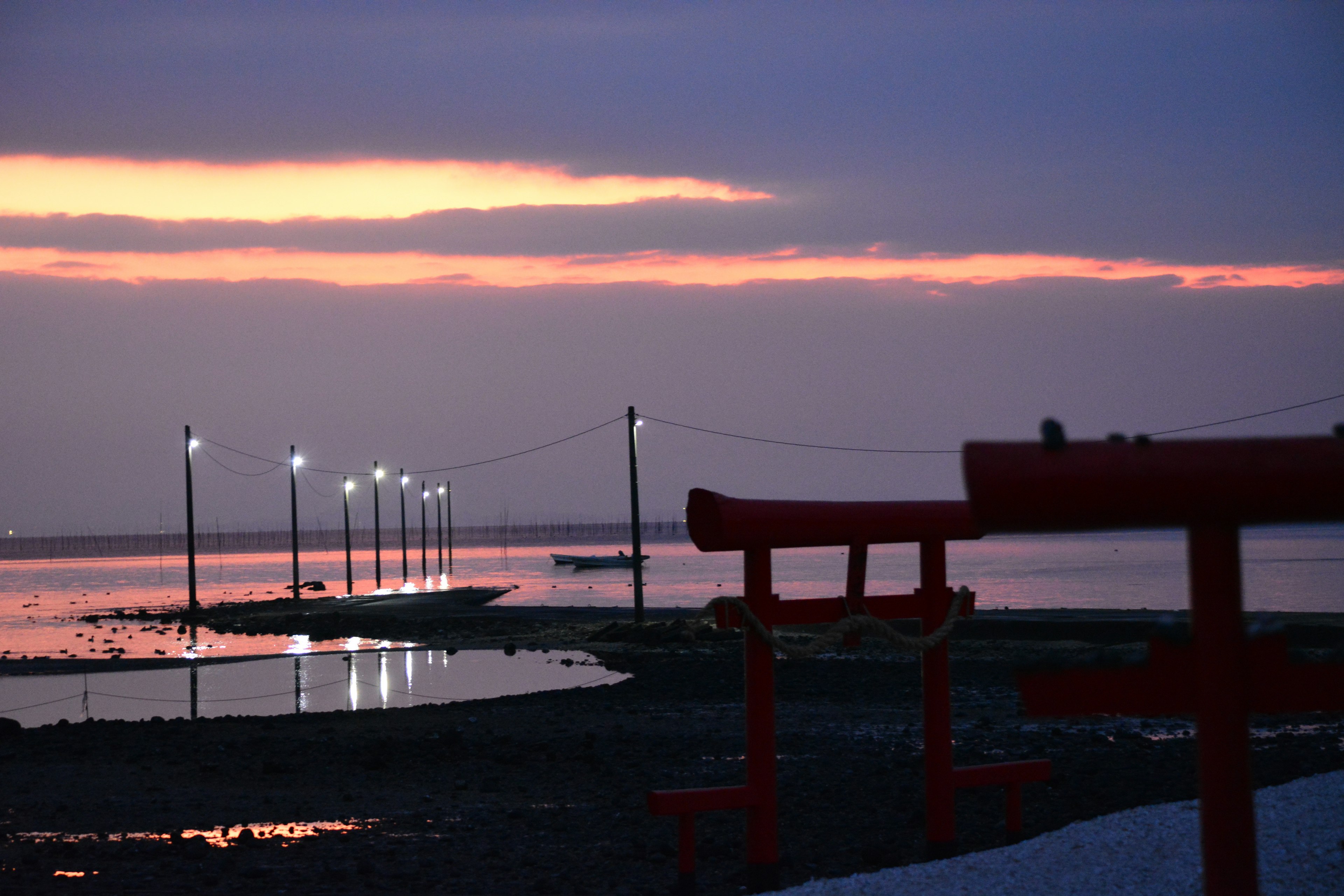 夕焼けの海に立つ赤い鳥居と灯りの並ぶ小道