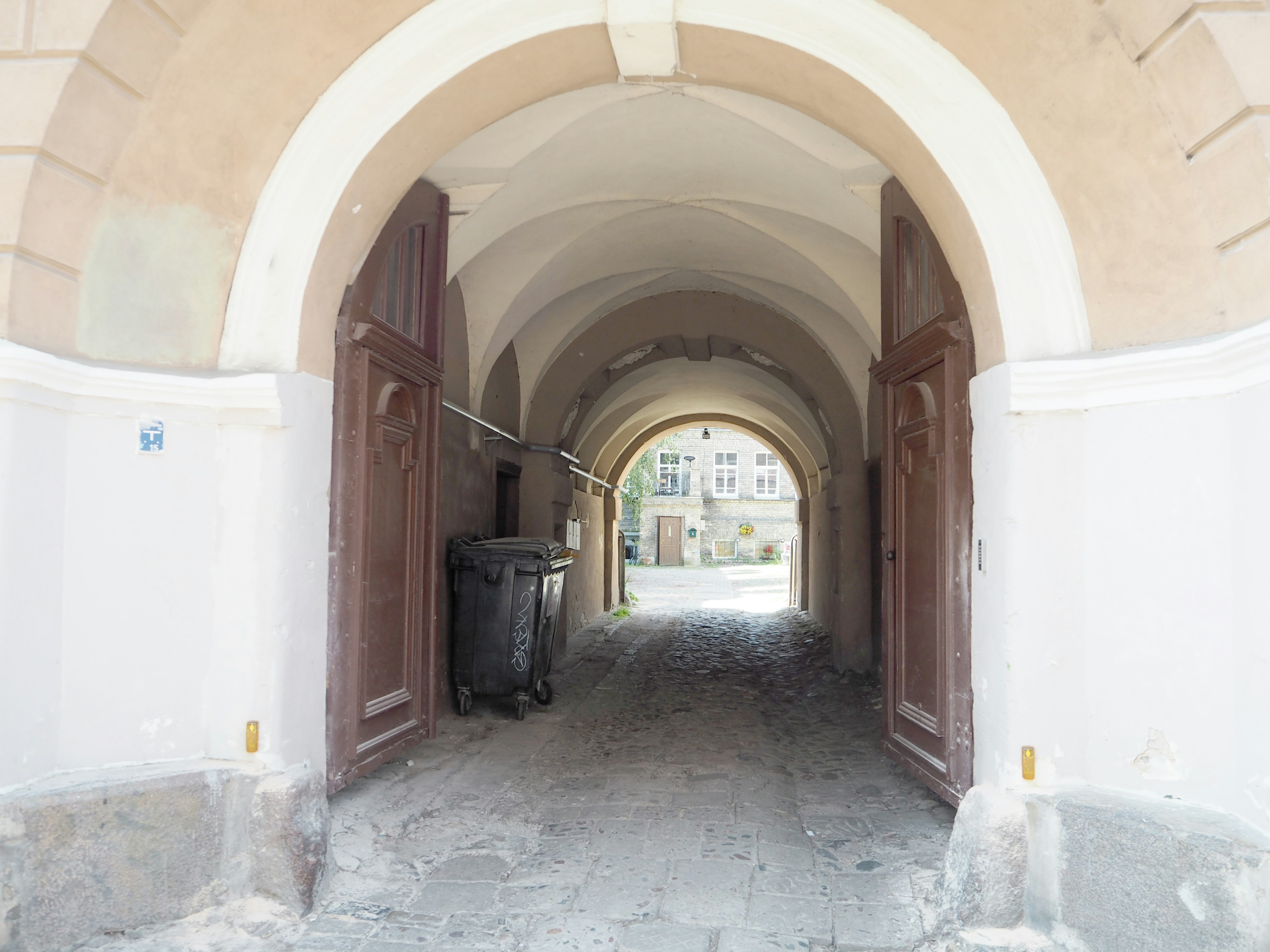 Archway leading to a pathway with old wooden doors and a trash bin