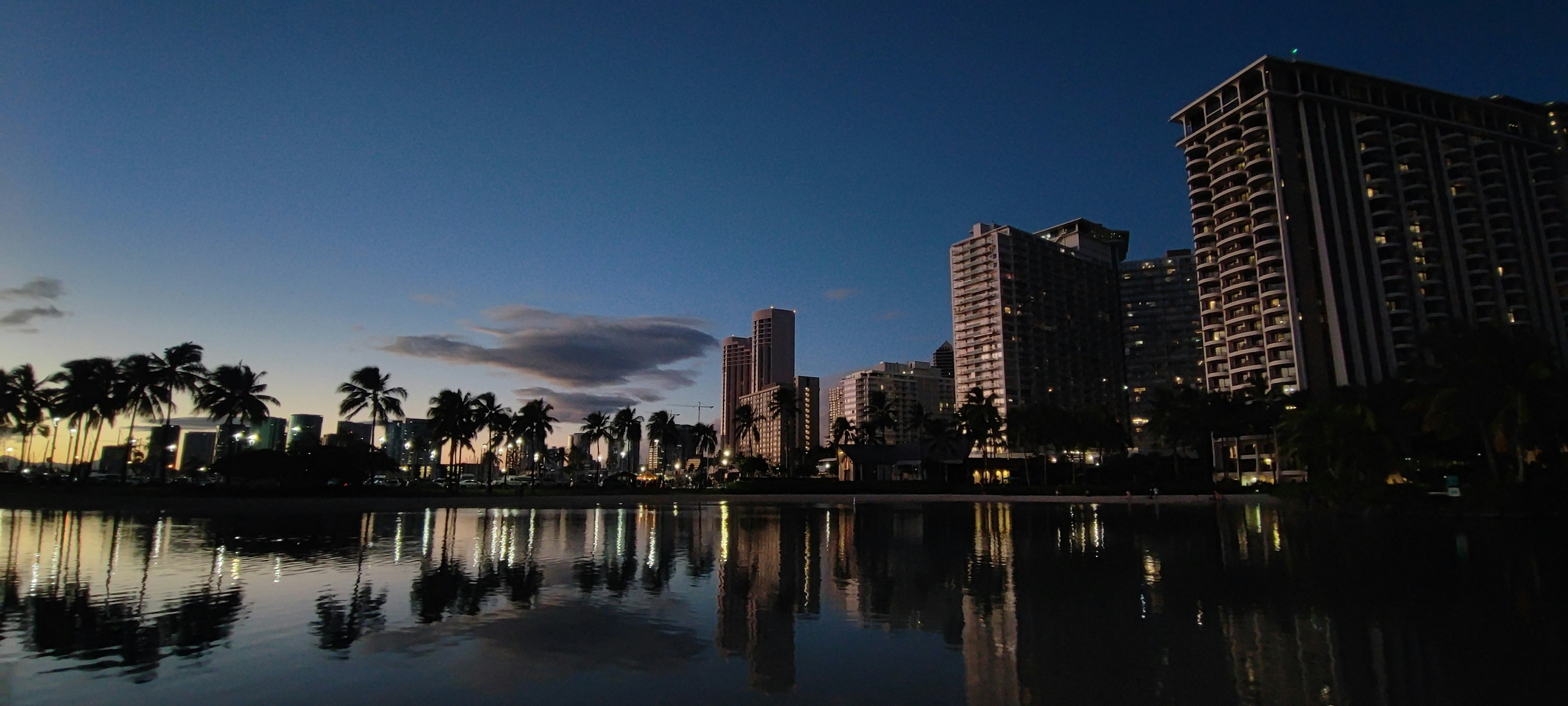 Vue nocturne d'une ville hawaïenne avec des silhouettes de bâtiments se reflétant sur une eau calme