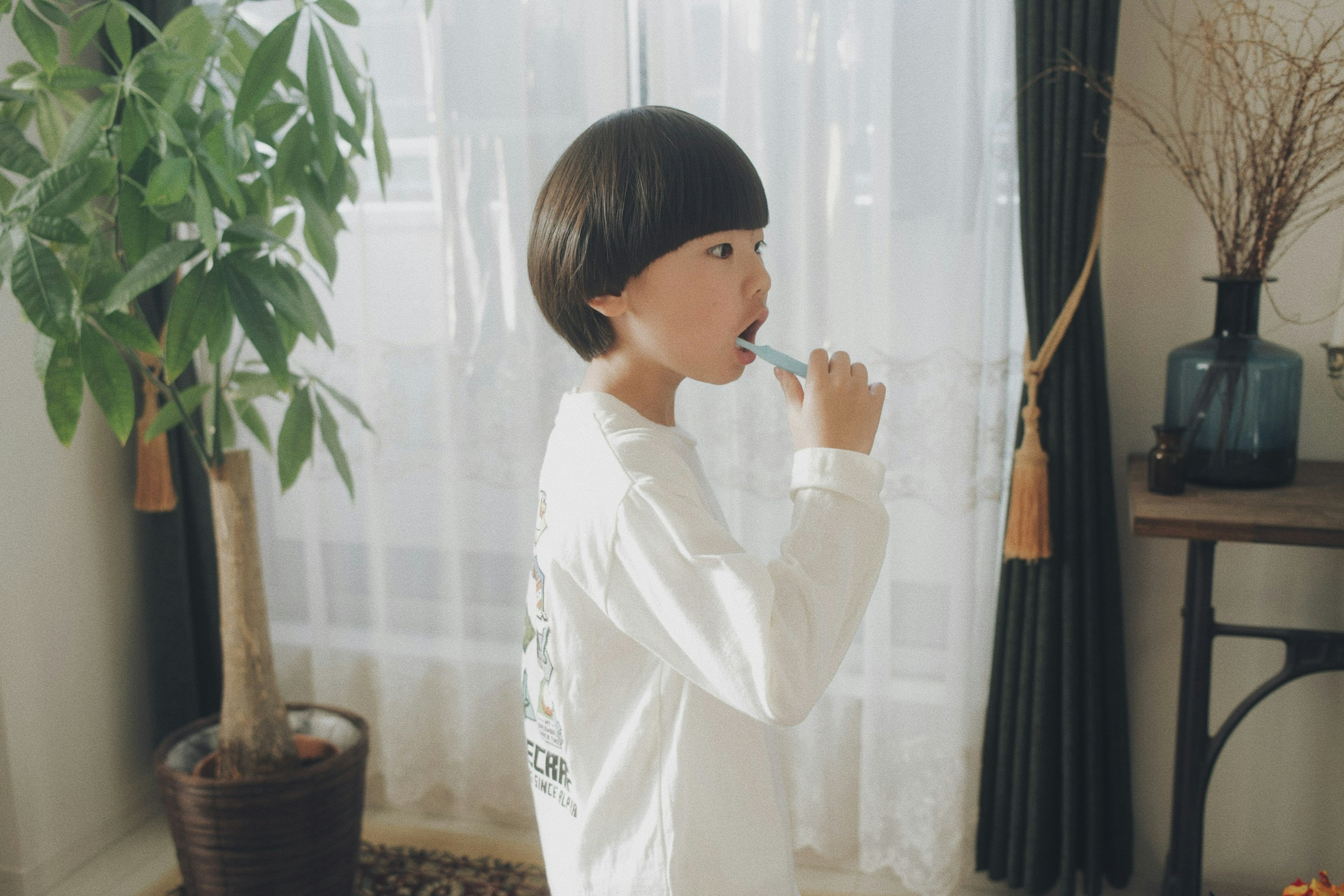 A child in a white shirt brushing their teeth by a window with a plant and decor visible