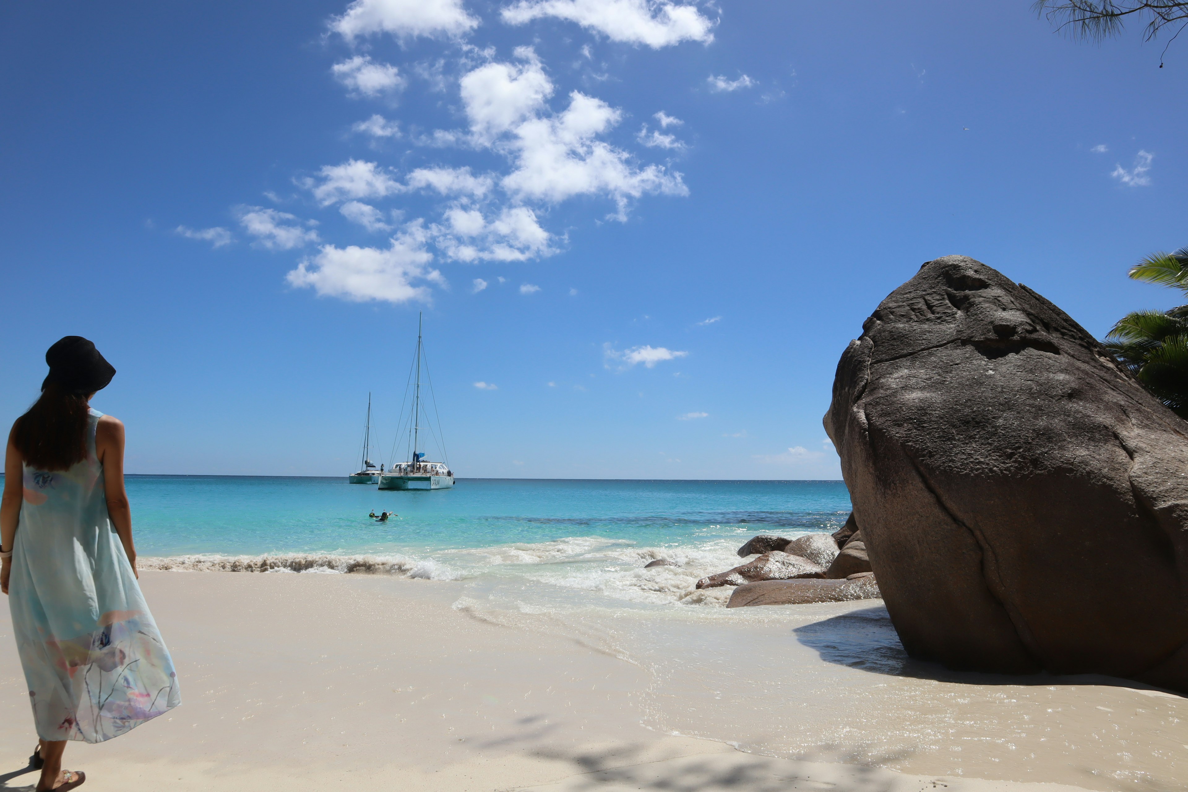 Donna che cammina su una bella spiaggia con acqua turchese e sabbia bianca, grande roccia nelle vicinanze, veliero in lontananza