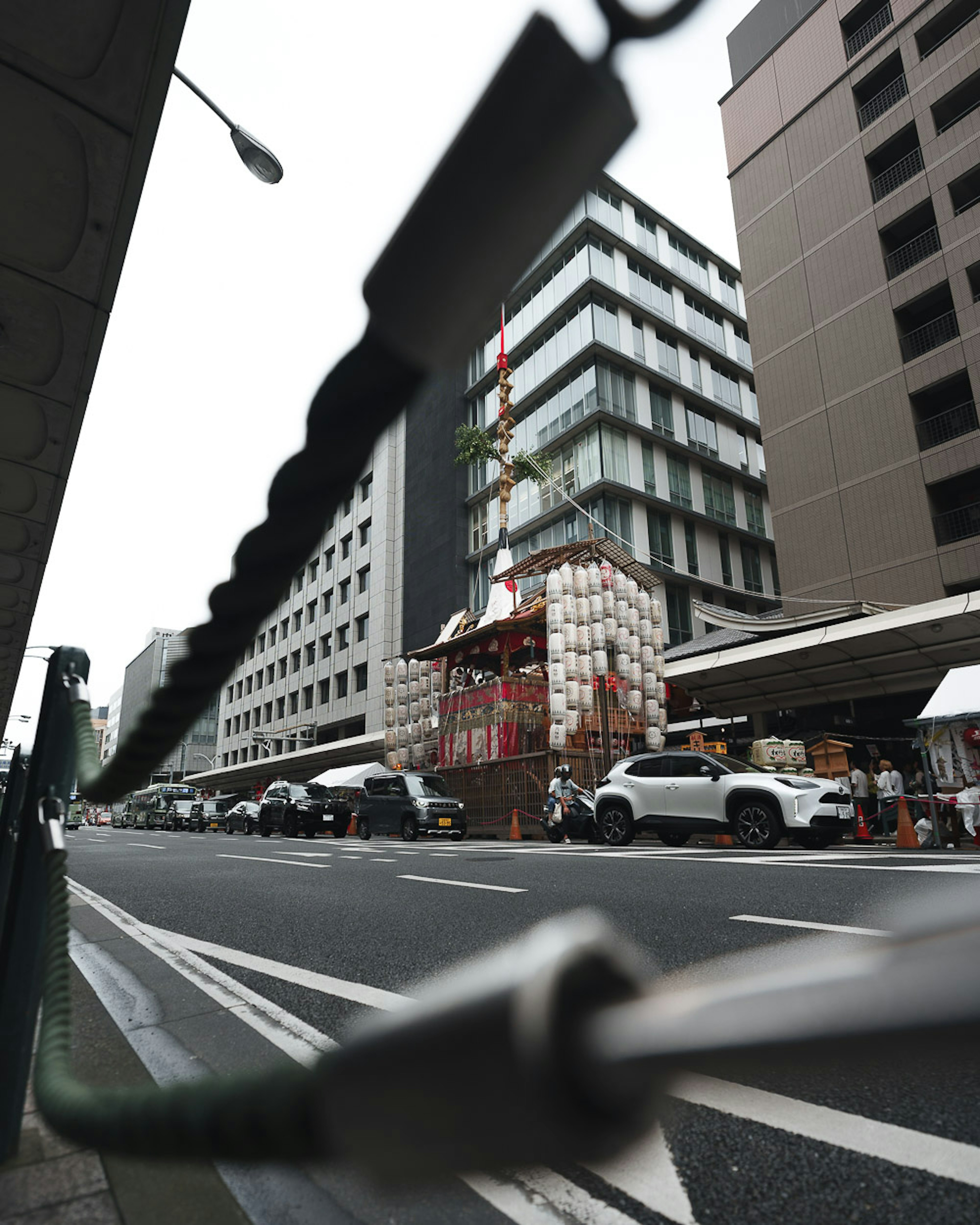 道路に面した建物と工事中の構造物があり、背後に高層ビルが立ち並ぶ