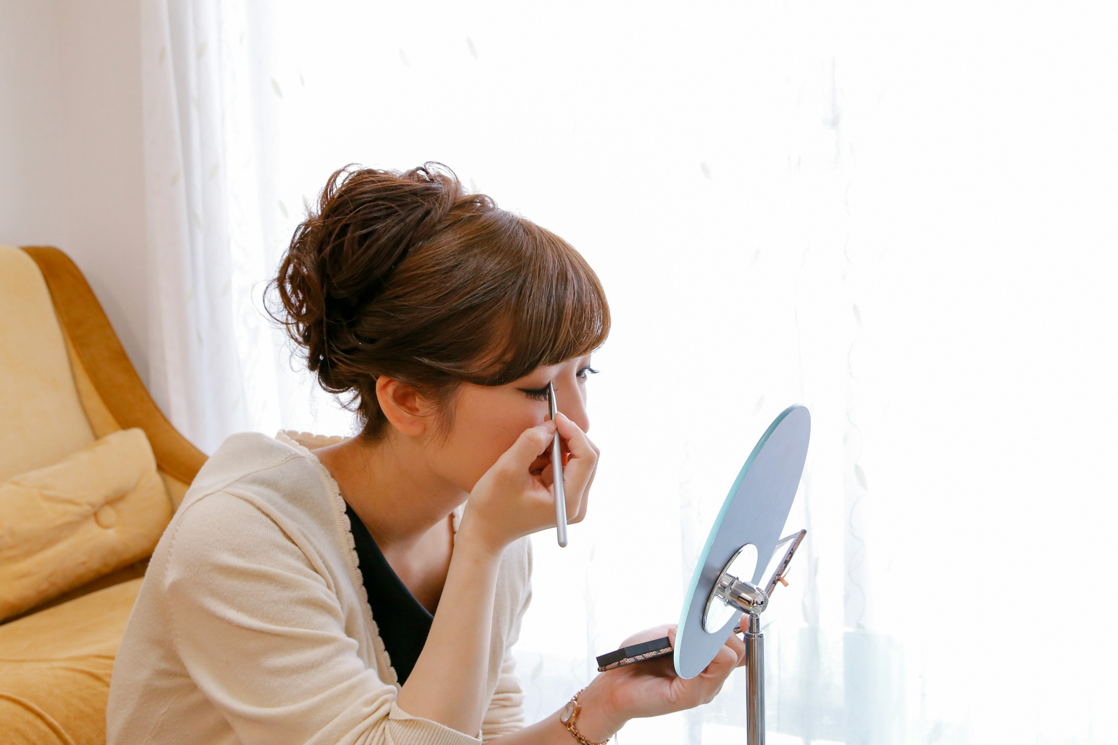 A woman applying makeup in front of a mirror in a bright room with natural light