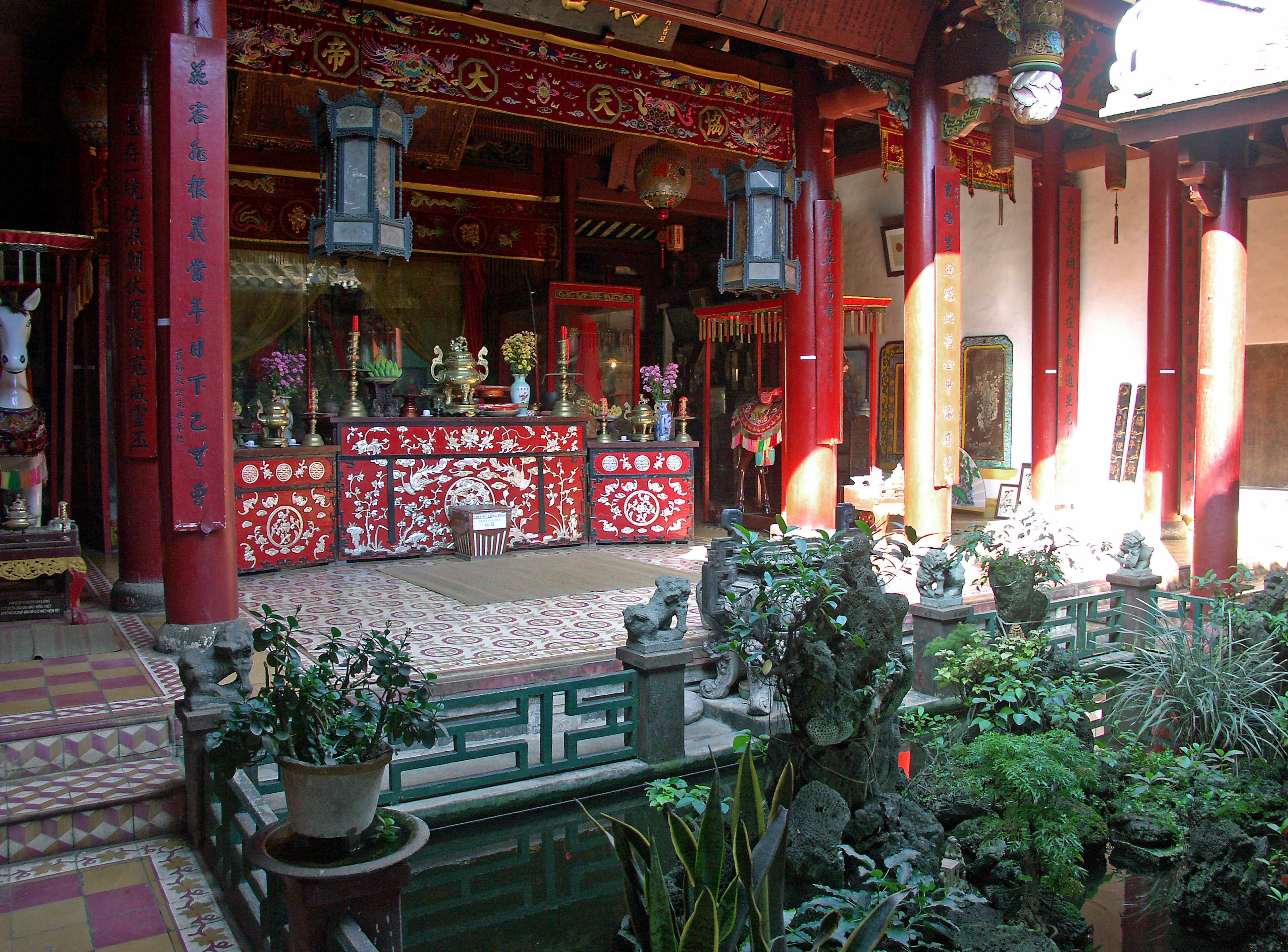 Vista interior de un templo chino tradicional con pilares rojos y decoraciones intrincadas, plantas y esculturas en un jardín sereno