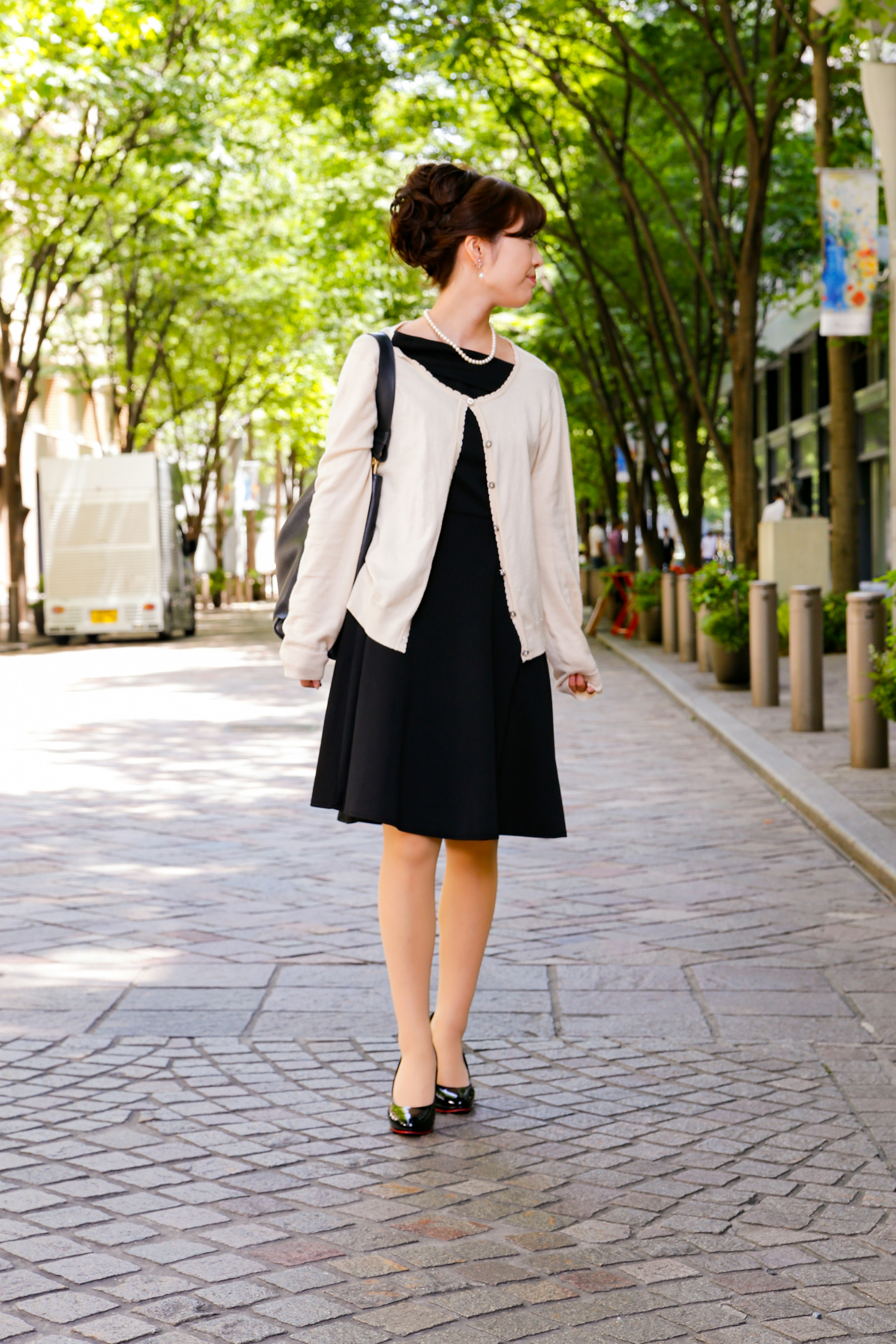 Une femme en robe noire et cardigan blanc marchant dans une ville avec des arbres verts