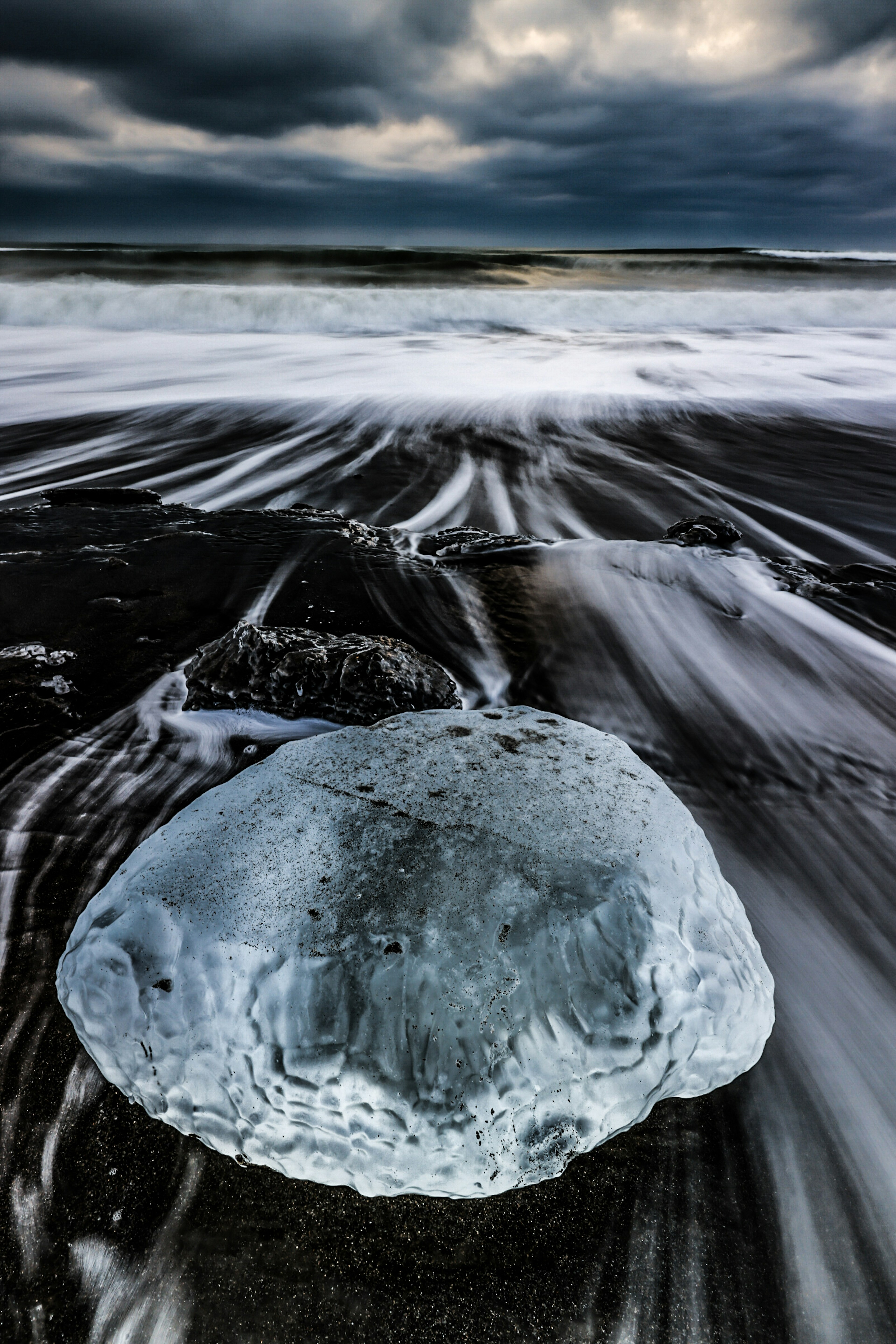 Ein blauer Eisblock auf einem schwarzen Sandstrand, der von Wellen gewaschen wird