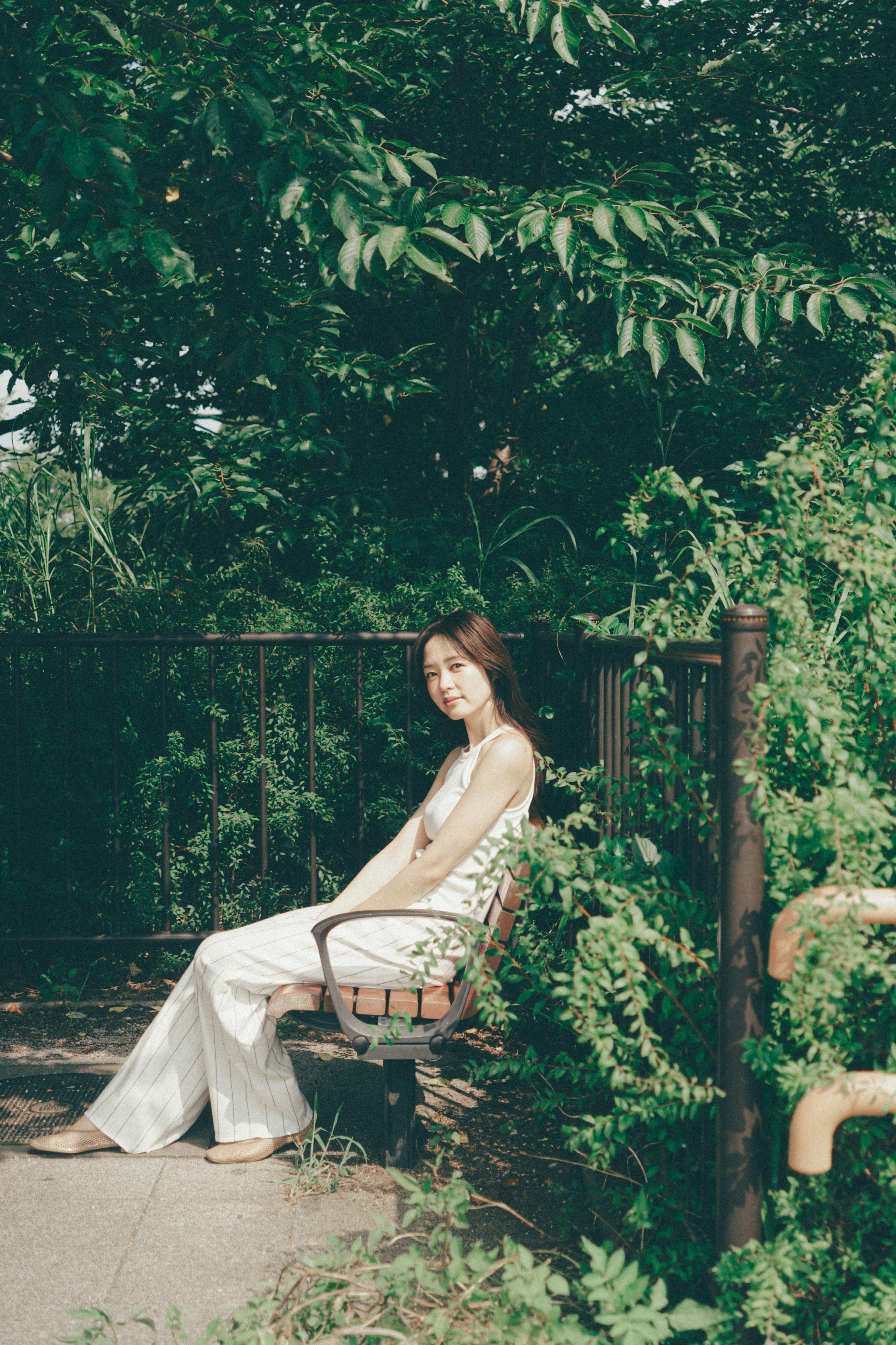 A woman sitting on a bench surrounded by greenery wearing a white outfit with a relaxed expression