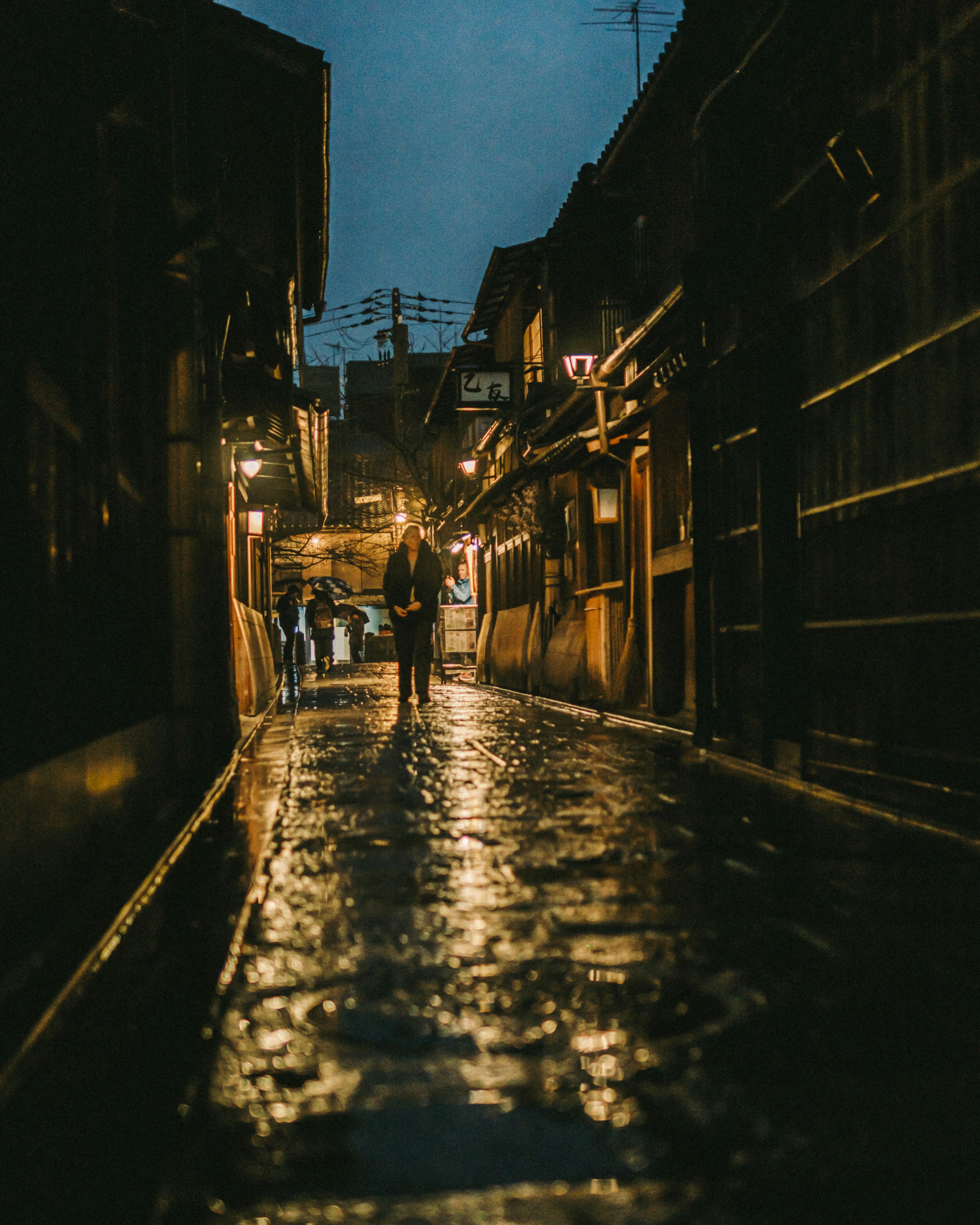 Eine Person steht in einer regennassen Gasse bei Nacht mit warmen Lichtern