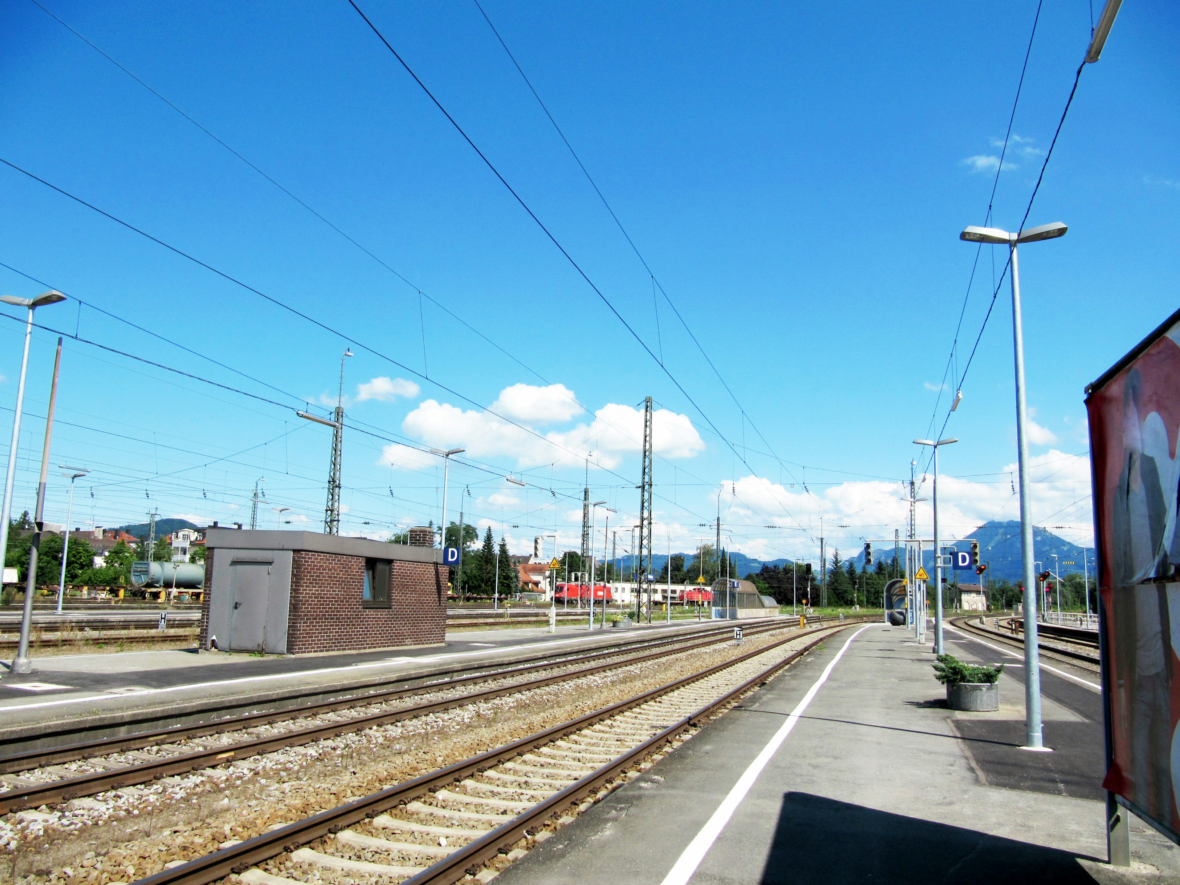 ภาพสถานีรถไฟใต้ท้องฟ้าสีฟ้าพร้อมเมฆขาวแสดงให้เห็นรางรถไฟและอาคารเล็ก