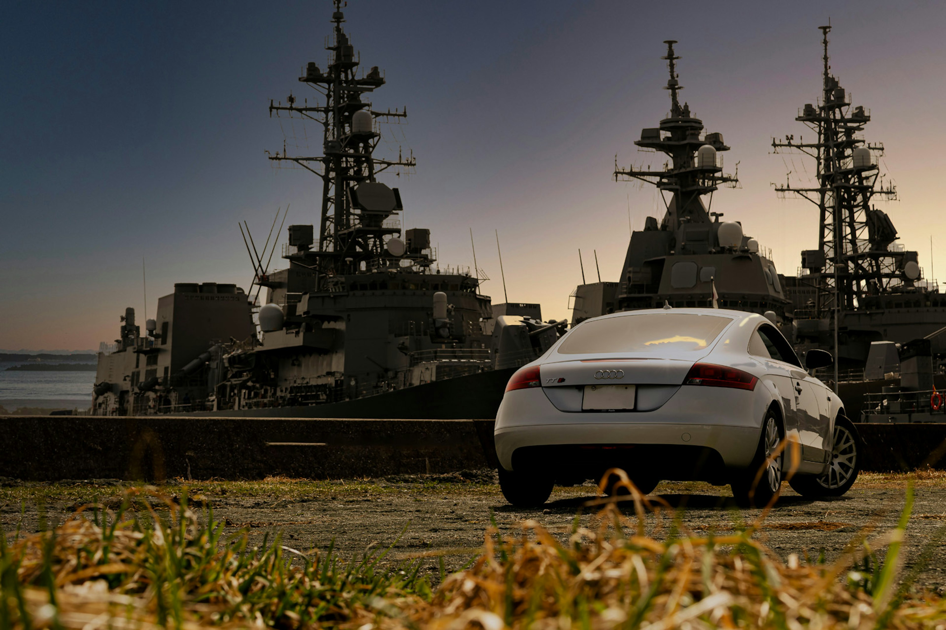 Coche deportivo blanco estacionado frente a barcos militares al atardecer