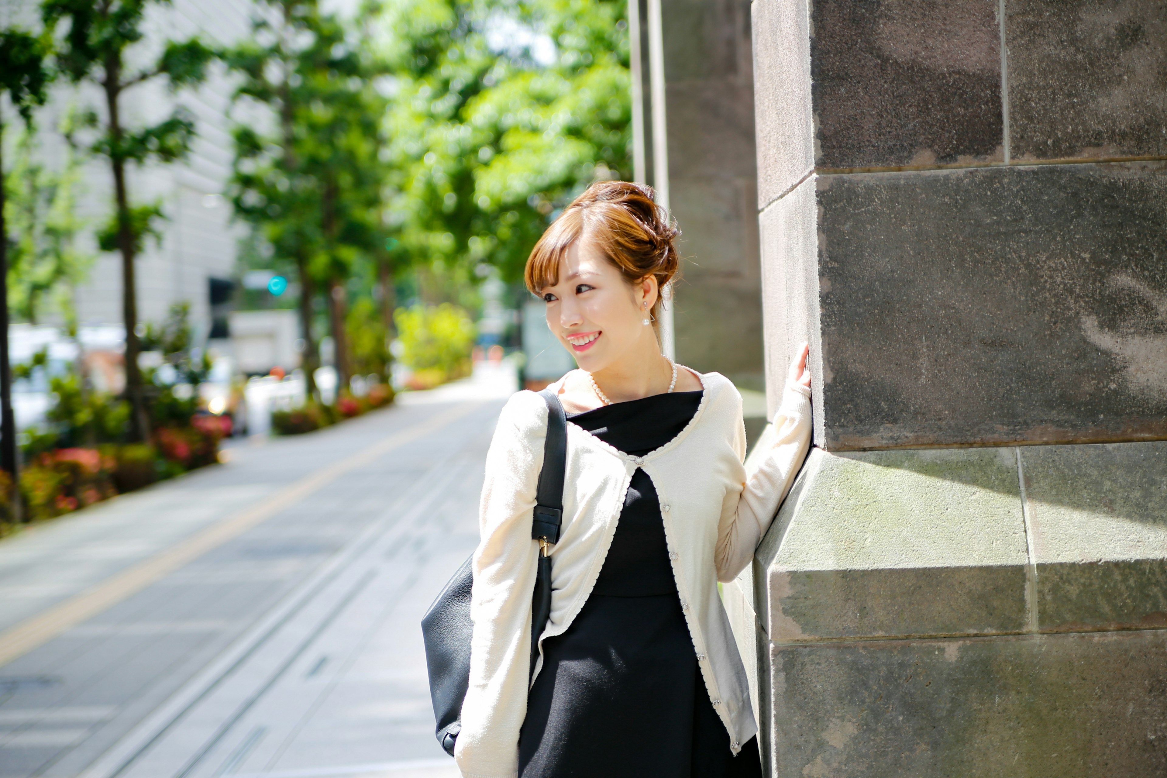 Femme debout dans la rue portant une robe noire et un cardigan blanc entourée de verdure et d'un mur en pierre