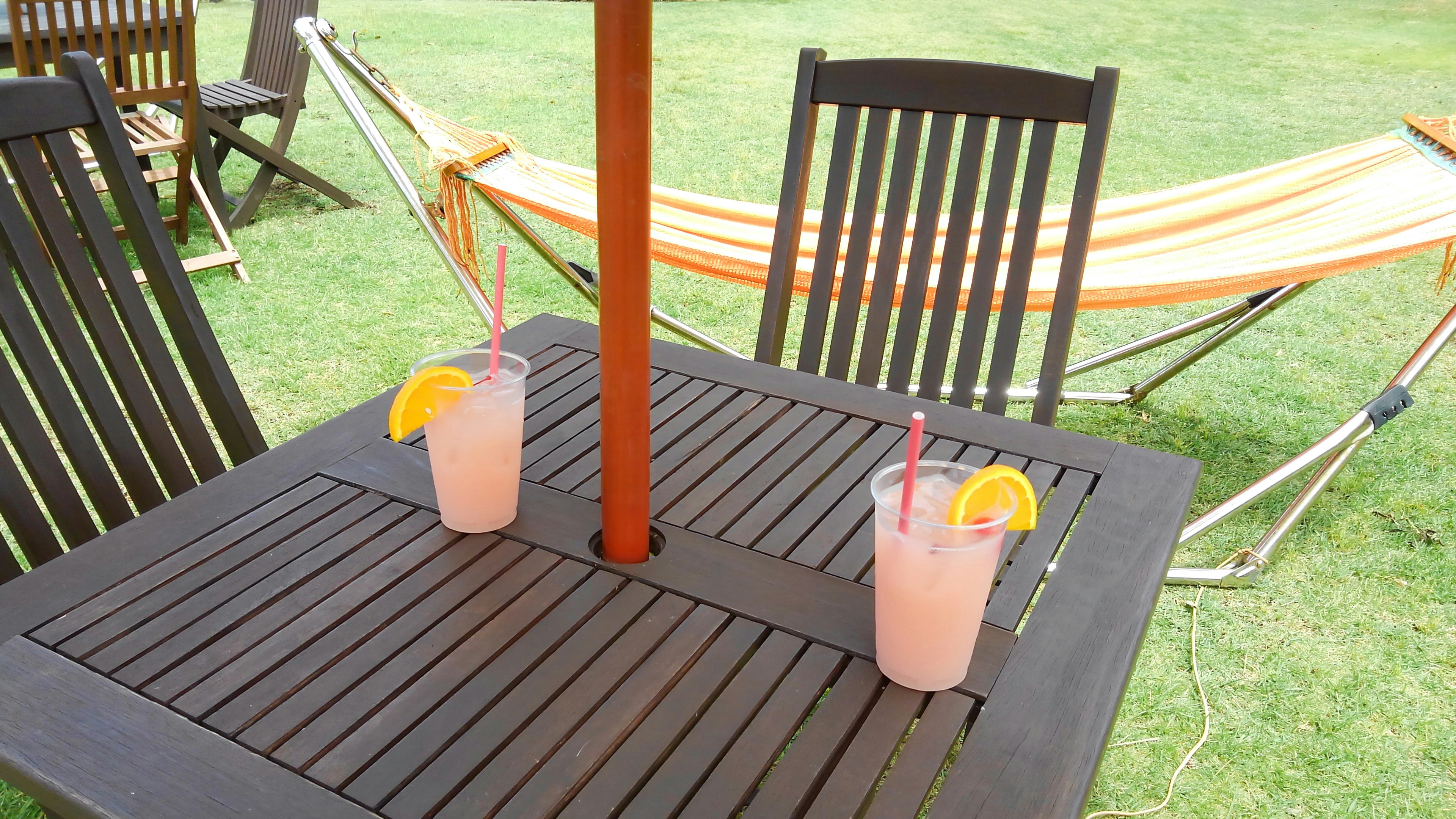 Two pink drinks with orange slices on a wooden table with chairs and a hammock in the background