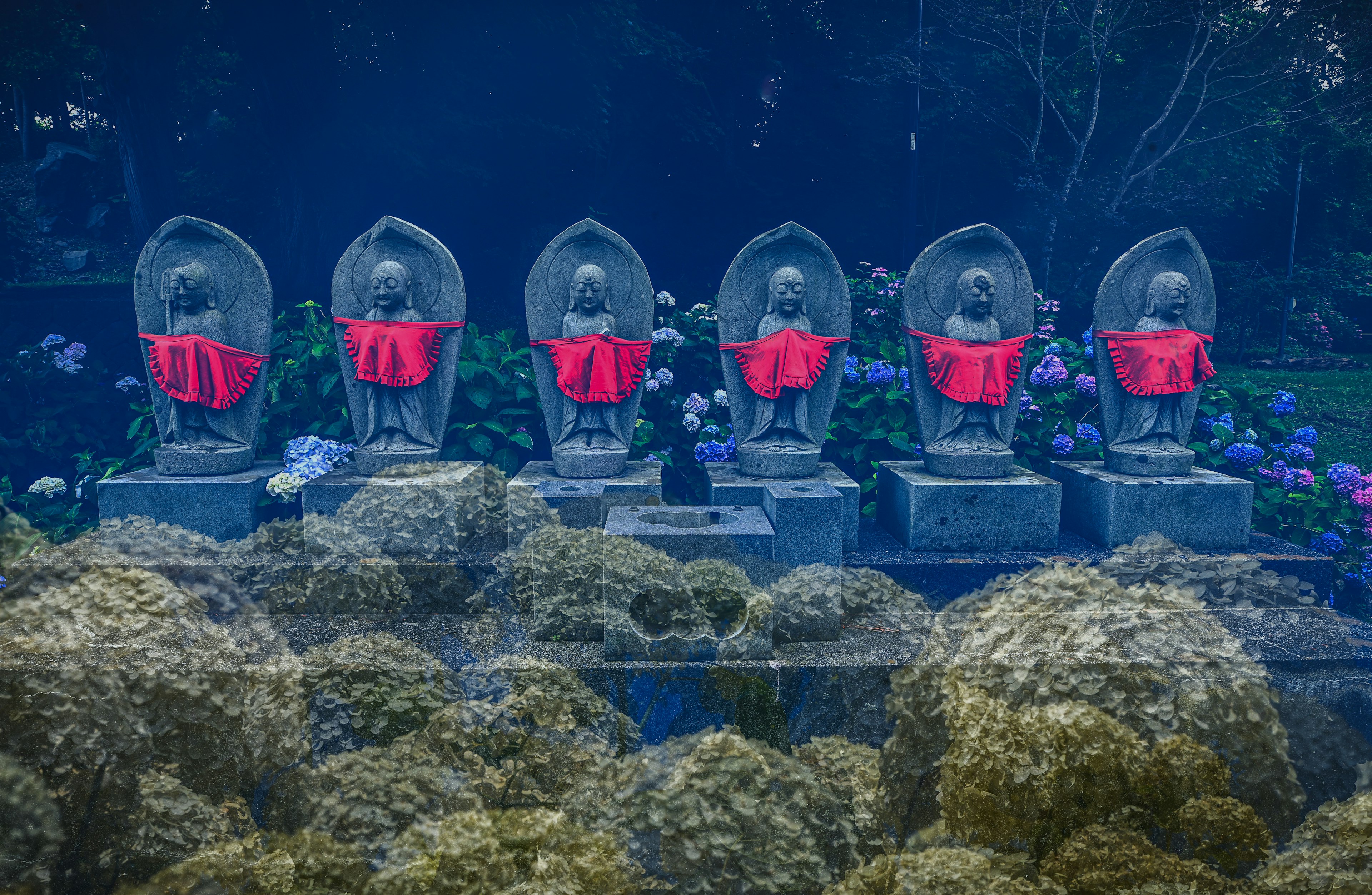 Six stone statues wearing red cloth in a garden setting