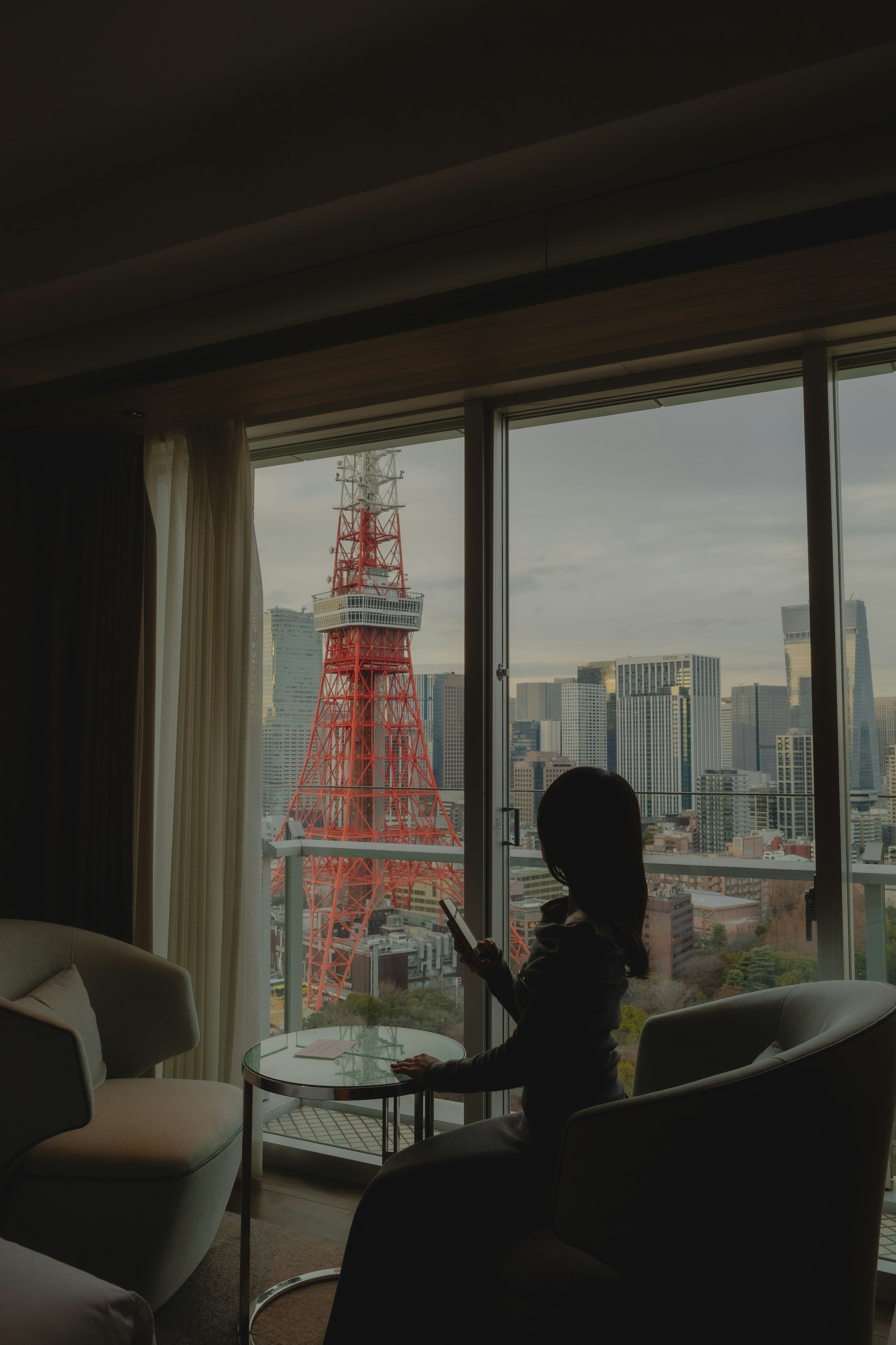 Seorang wanita duduk di dalam kamar hotel dengan pemandangan Menara Tokyo melalui jendela