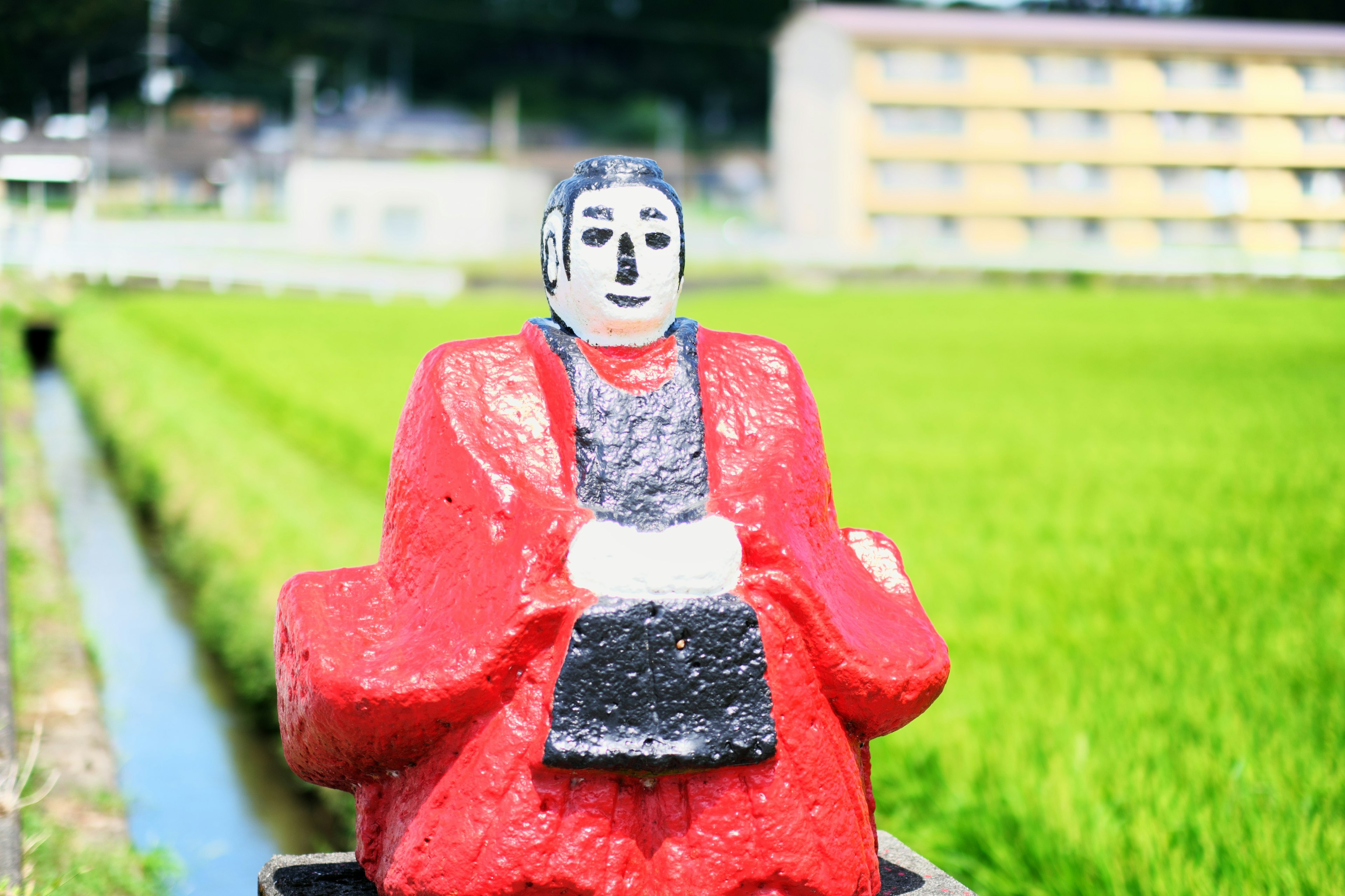 A red-clad statue of a Buddha figure near a rice field