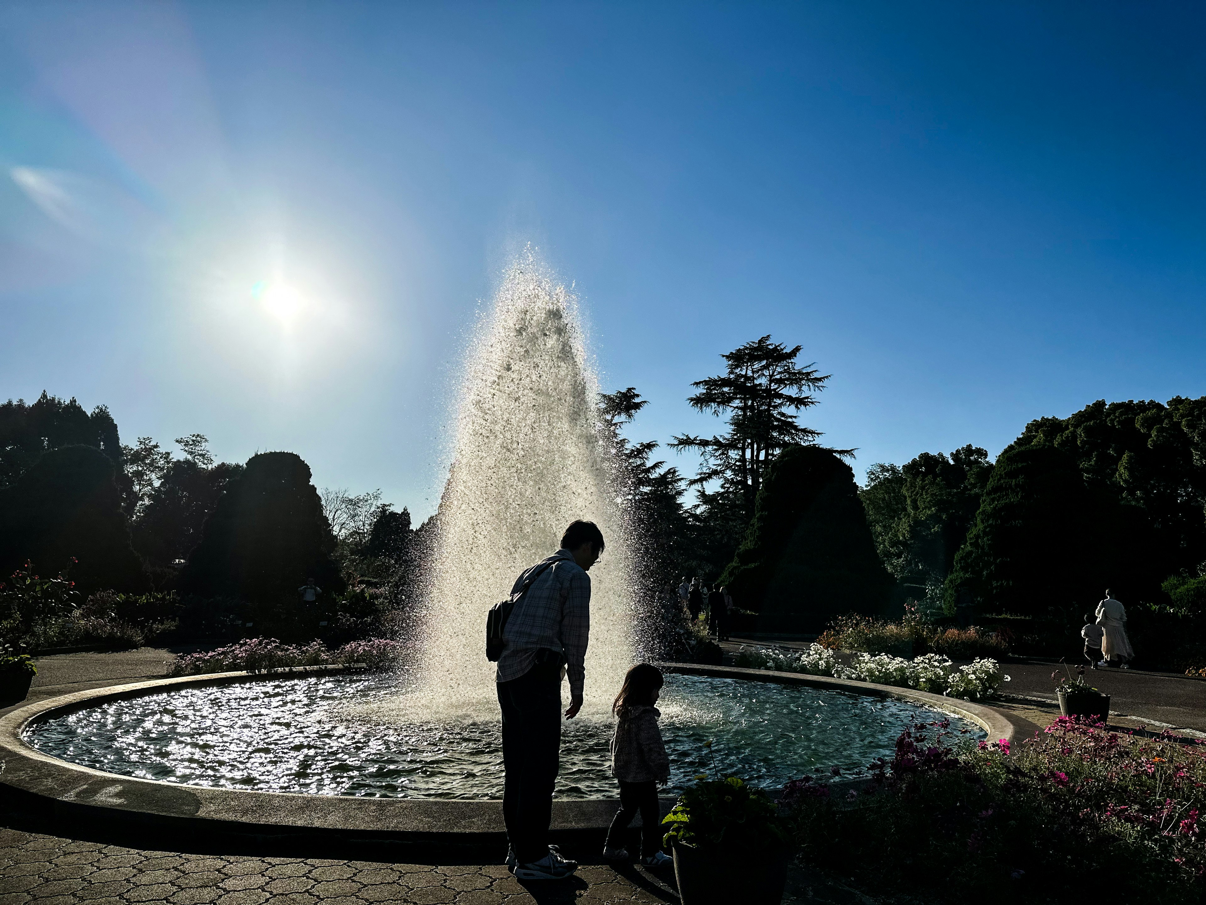 Silhouette di un genitore e un bambino davanti a una fontana in un parco con cielo blu