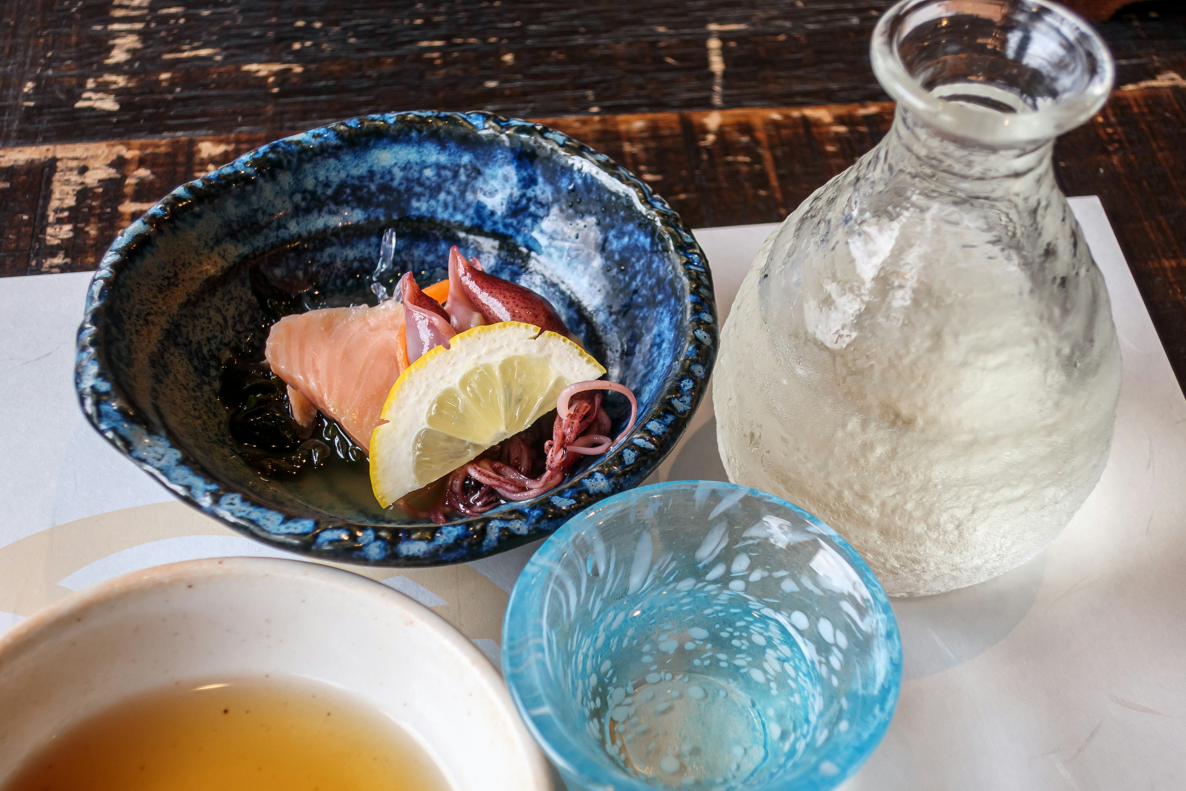 A blue dish with lemon and prosciutto alongside a clear bottle of beverage on a wooden table