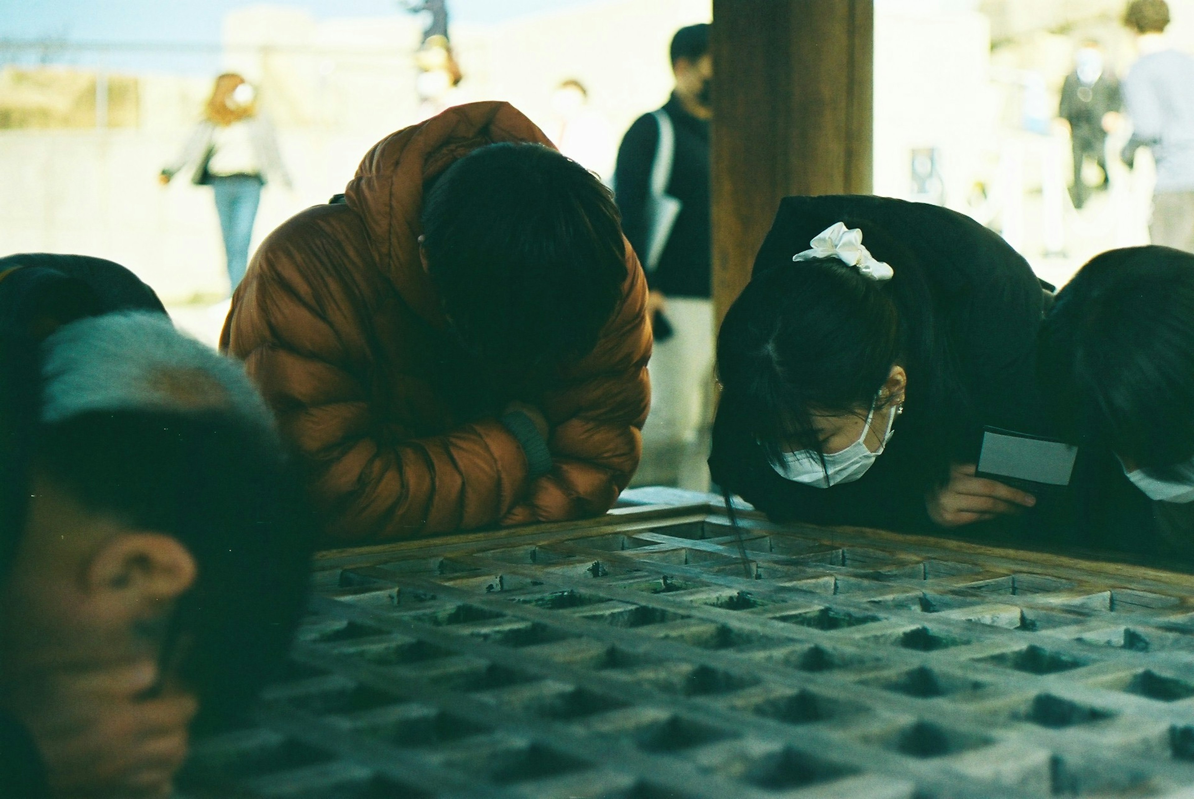 Children with heads down near a textured surface and people in background