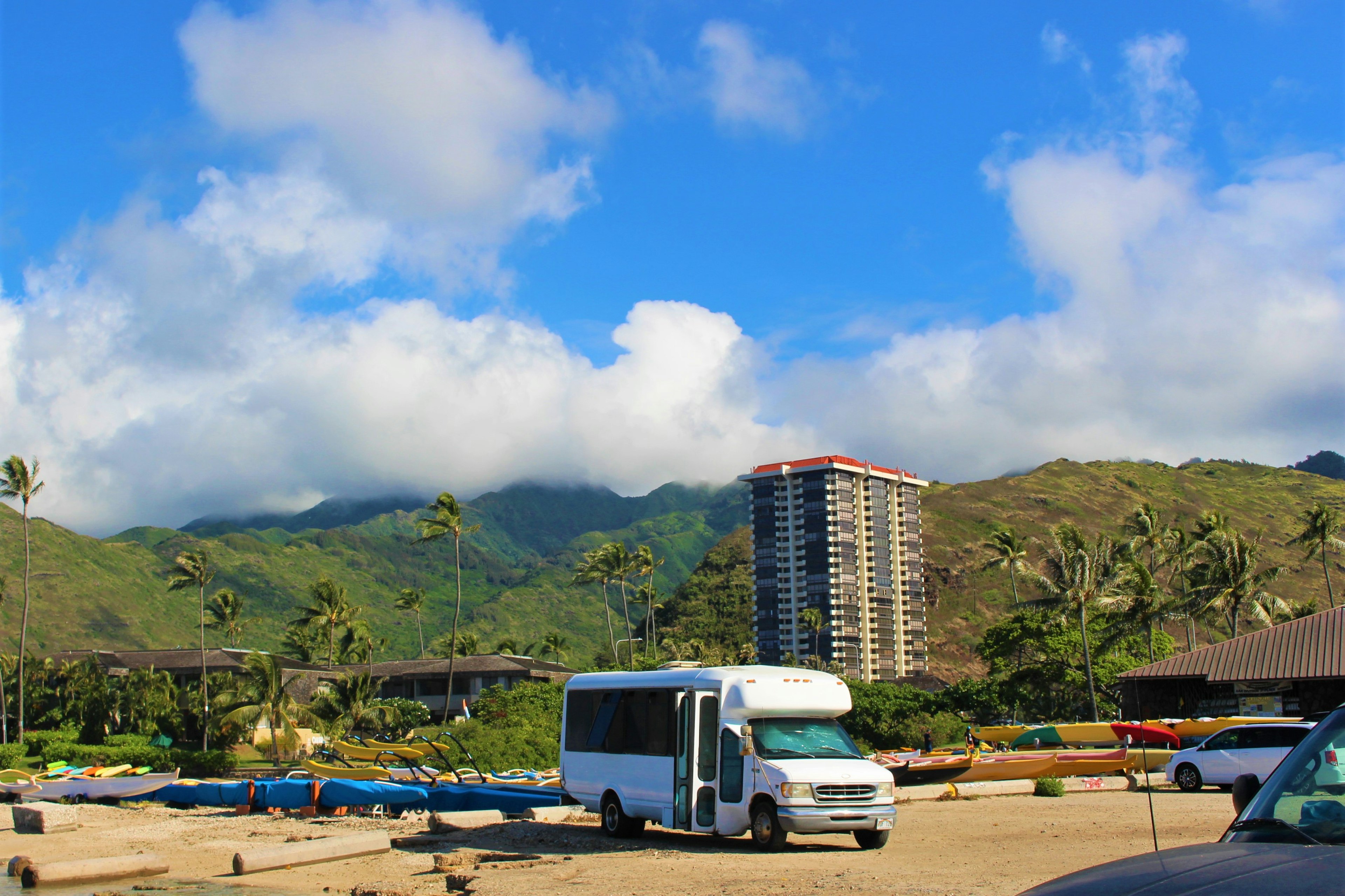 A scenic view with a white bus and a high-rise building under a blue sky featuring mountains and palm trees in the background