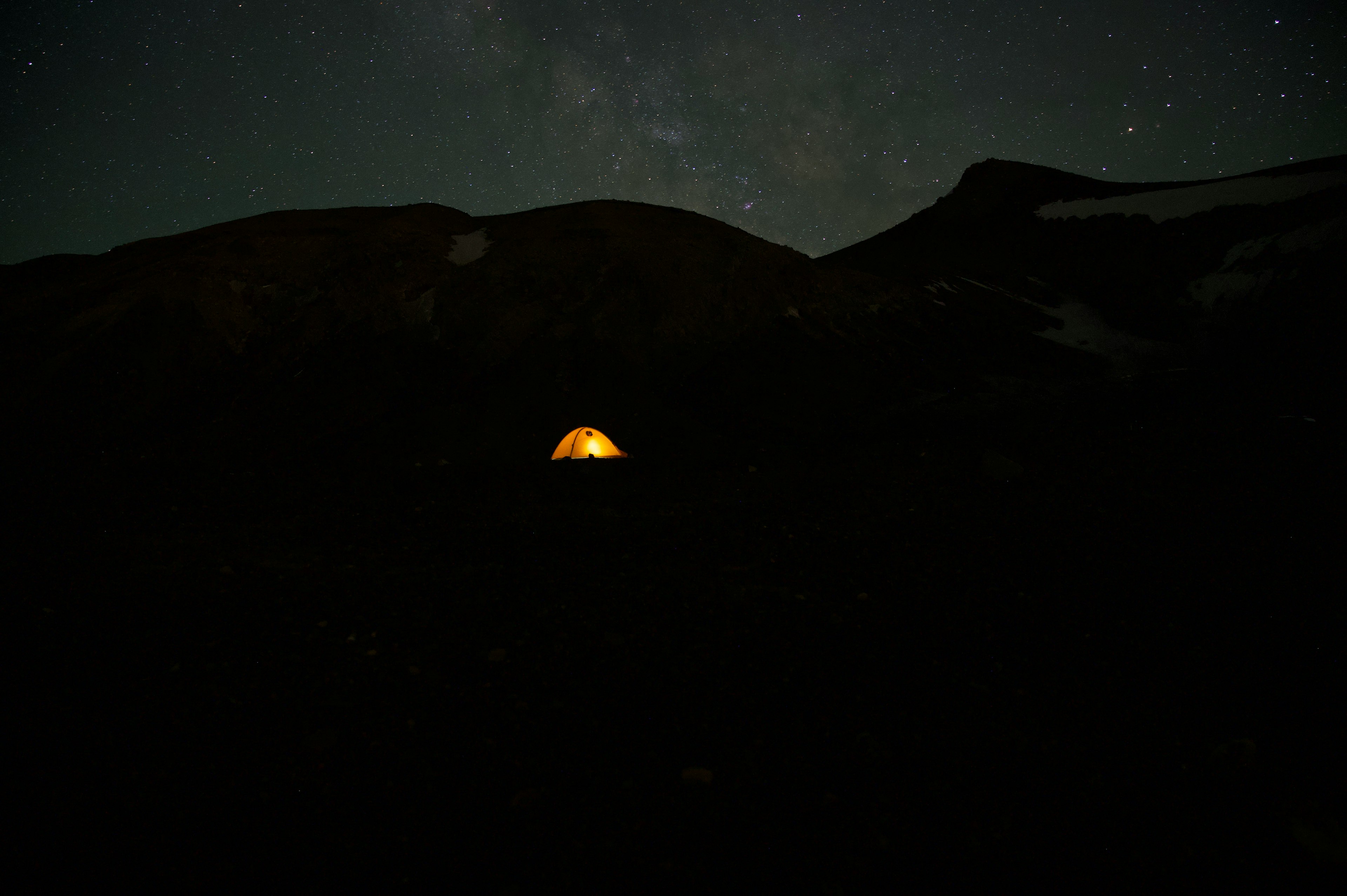 Sebuah tenda kecil bersinar dalam kegelapan di bawah langit berbintang dengan siluet gunung